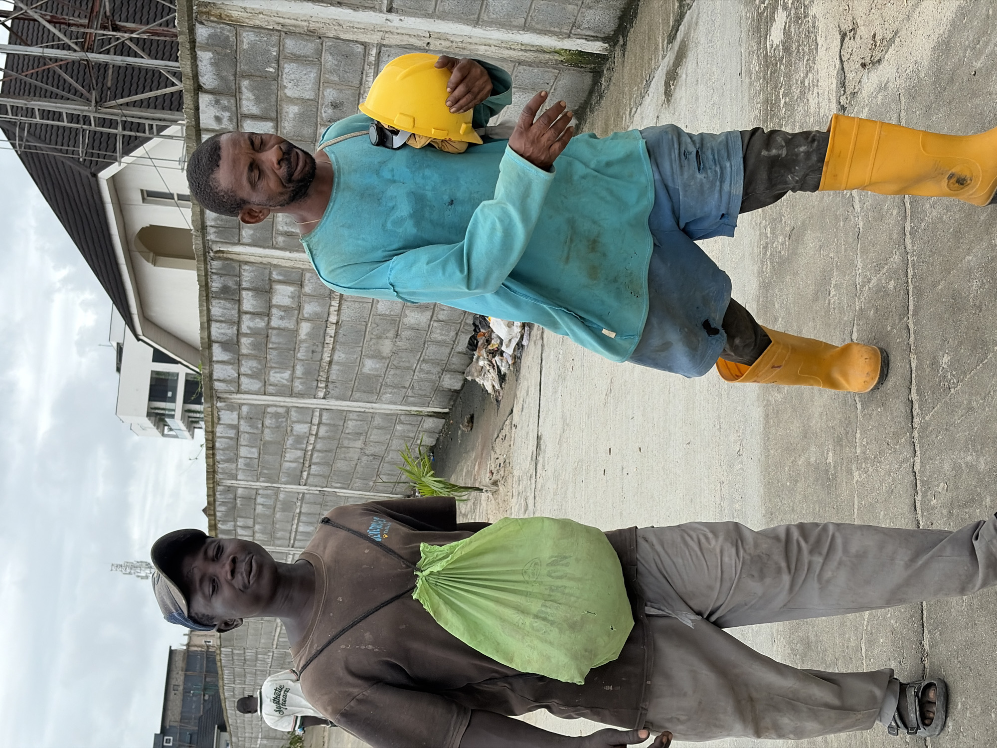 Two men are walking on a paved surface in Alaguntan, Nigeria. The man on the left, wearing a dark t-shirt and grey pants, carries a light green drawstring bag over his shoulder. He is wearing a dark baseball cap and sandals. The man on the right, dressed in a teal long-sleeved shirt and blue shorts, holds a yellow hard hat. He is wearing dark pants underneath his shorts and bright yellow rubber boots. The background features a concrete block wall, some buildings, and an overcast sky. Debris is visible along the wall.