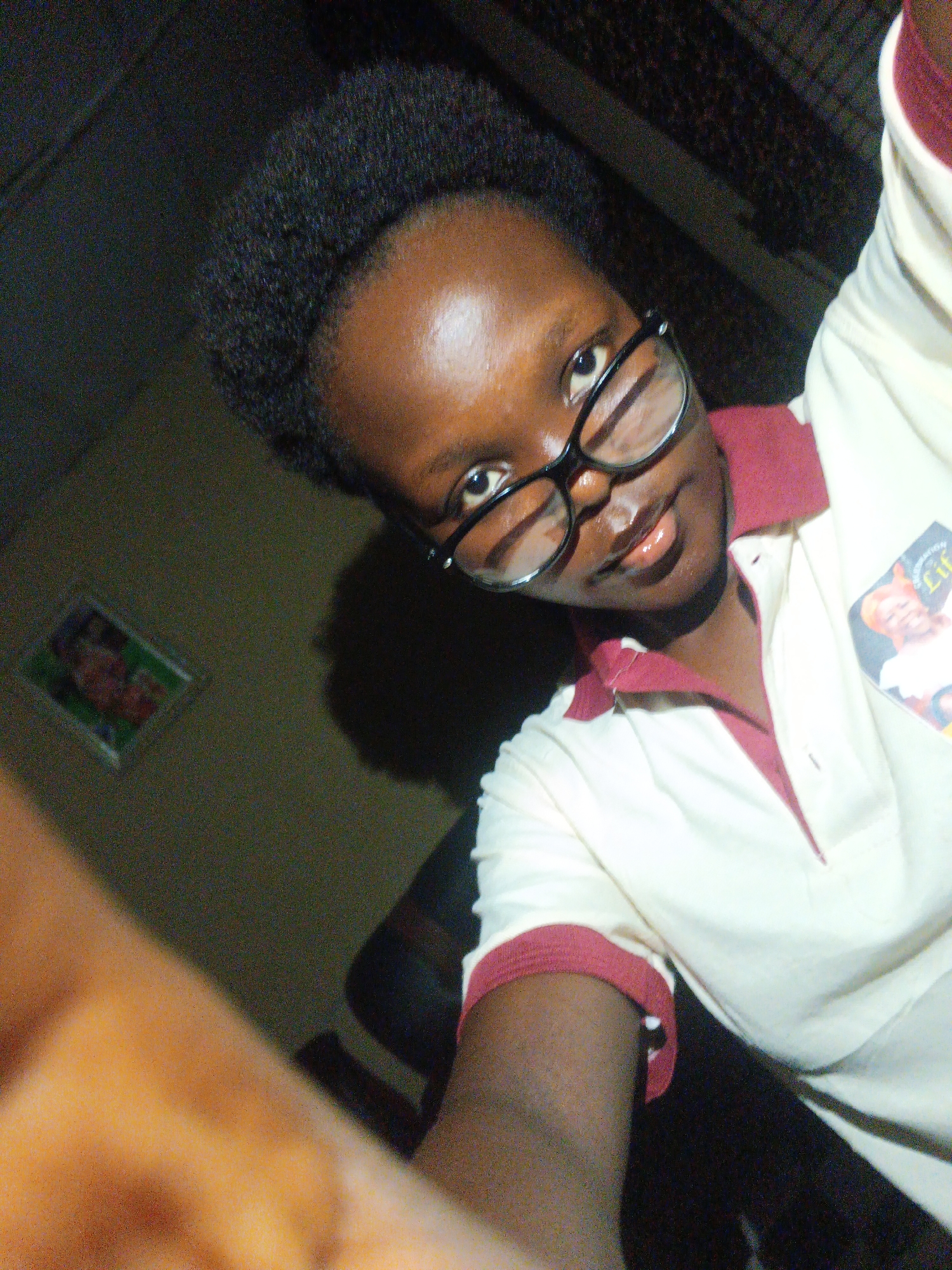 A dark-skinned person is taking a selfie indoors. They are wearing glasses with black frames and a collared shirt that is white with maroon trim on the collar and sleeves. There is a sticker or patch on the shirt. The person has short, dark hair. There is a picture hanging on the wall in the background and a chair is also visible. The specific location within Onitsha, Nigeria, cannot be determined from the image.