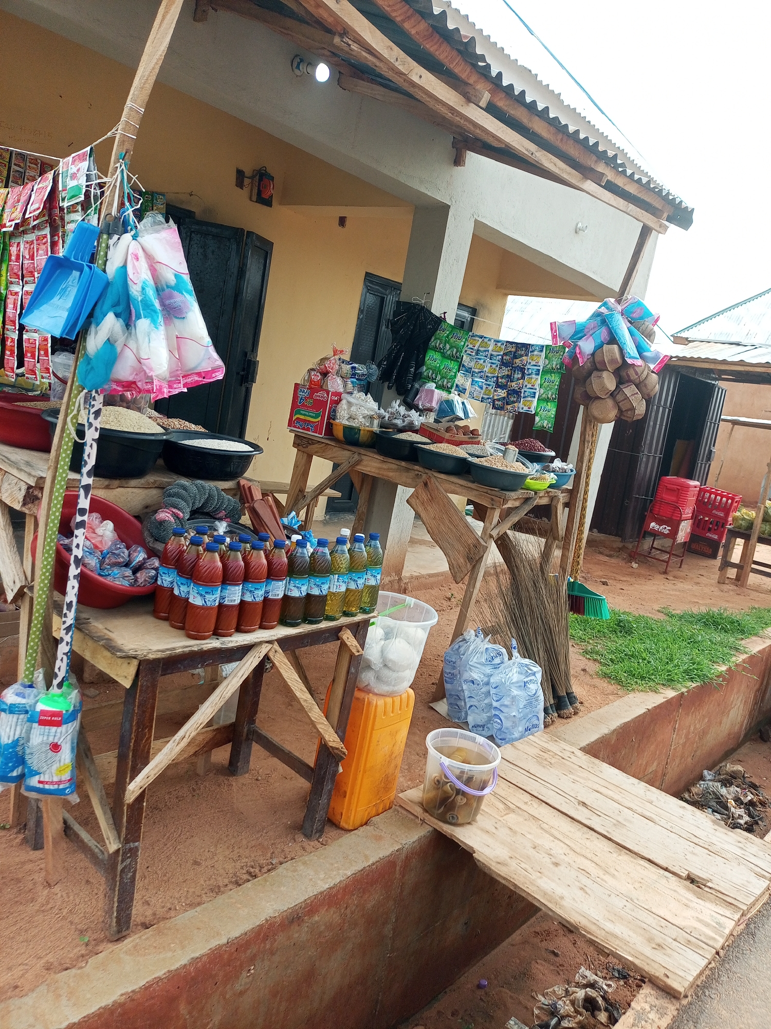 A market stall is set up outdoors in Tempure, Nigeria. The stall is constructed from wood and corrugated metal roofing. Several tables are laden with various goods, including bags of rice, bottles of oil, and packaged food items. In the foreground, a makeshift wooden bridge spans a ditch filled with debris. Several brooms are leaning against a table. A bright orange plastic container is visible near the right side of the frame, and a clear plastic bucket containing spools of purple thread sits on the wooden bridge. In the background, a building with yellow walls and a dark door is partially visible. The ground is covered in sandy soil.