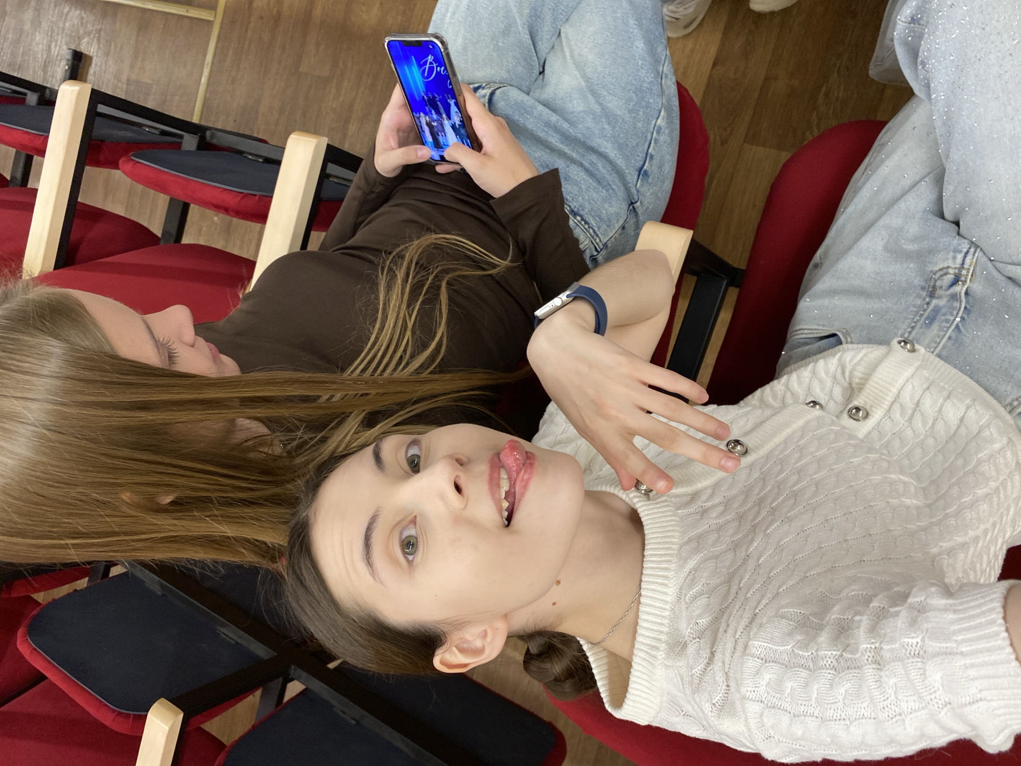 Two young women are seated in an indoor space, likely a theater or auditorium in Votkinsk, Russia, given the arrangement of red and black seating. The woman closer to the camera is lying back, sticking her tongue out, and wearing a white sweater and light blue jeans. The other woman is seated upright, looking at her smartphone, wearing a brown top and light blue jeans. Several empty seats are visible in the background. The floor is made of wooden planks.