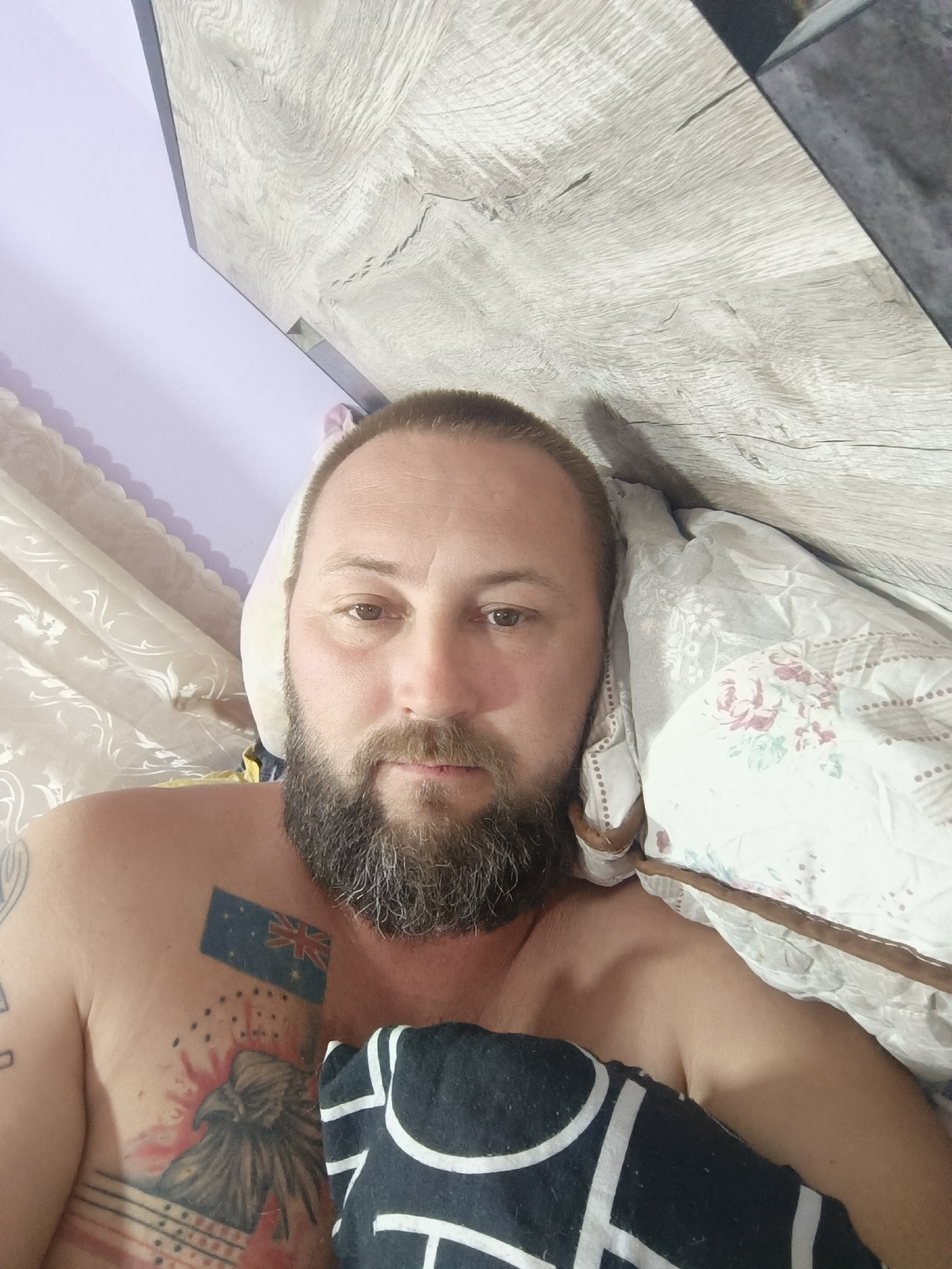The media file shows a close-up, overhead shot of a man lying in bed, presumably in a bedroom. The man has a beard and short hair. He is shirtless, revealing tattoos on his chest, including what appears to be an Australian flag and an eagle. He is partially covered by a pillow with a floral pattern and a dark blanket with white geometric designs. The background shows a wooden headboard and a wall with purple and white curtains. The lighting suggests it is daytime. The man's expression is neutral, and he appears to be looking at the camera. There are no visible text elements in the image. The general context provided states the location as "Taraz city administration, Kazakhstan," however, this location context is not evident from the visual information within the image itself.