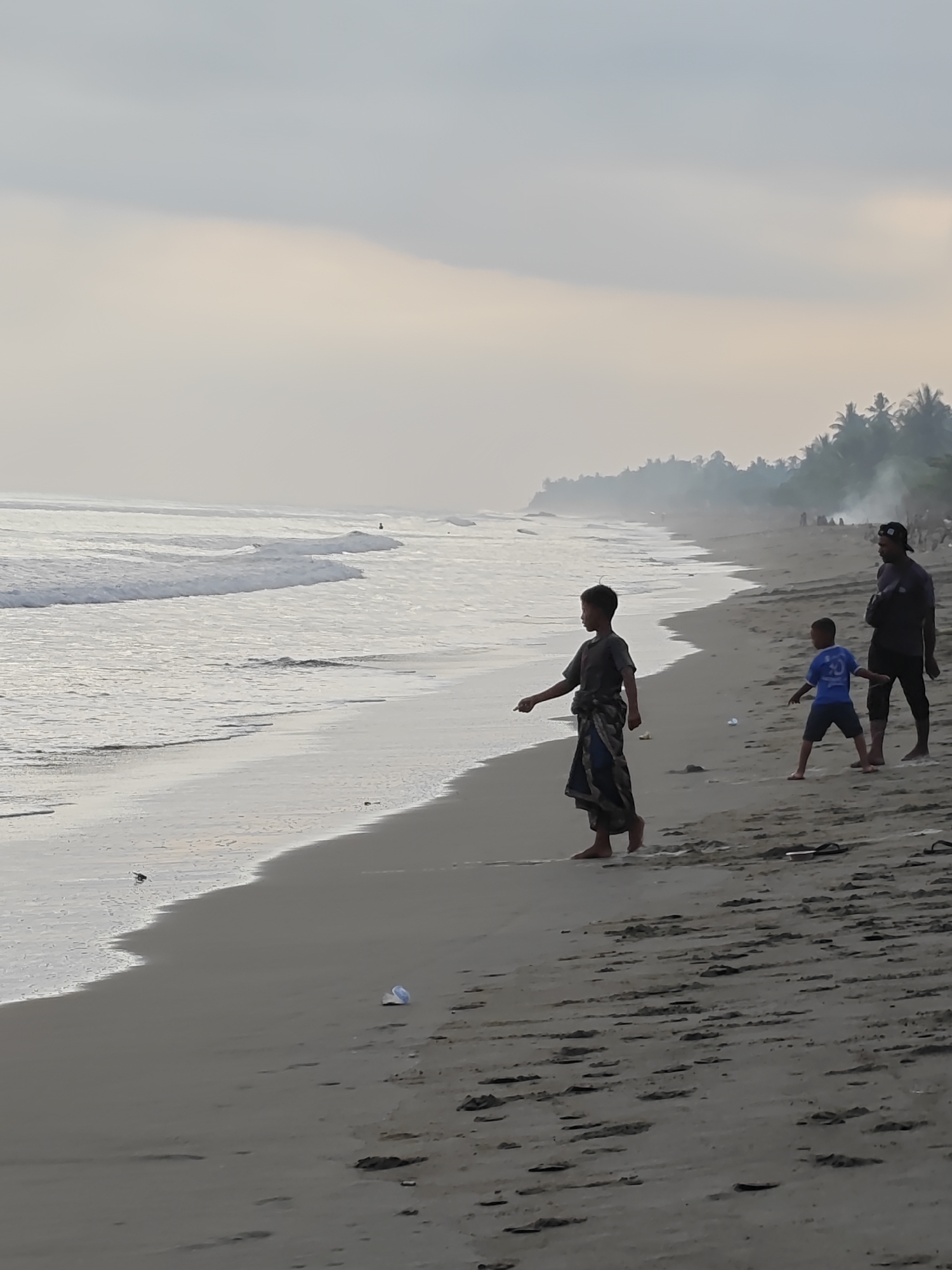 A group of three people, two children and one adult male, stand on the shore of a beach in Curehtunong, Indonesia. The children are in the foreground, with one child wearing a sarong and the other wearing a blue t-shirt. The adult male stands behind them, wearing a dark shirt and a baseball cap. The ocean is to the left of the frame, with waves gently rolling onto the shore. In the background, a line of palm trees and smoke suggest a village or settlement along the coastline. The sky is overcast, with soft light filtering through the clouds. The overall scene depicts a serene moment on a tropical beach.