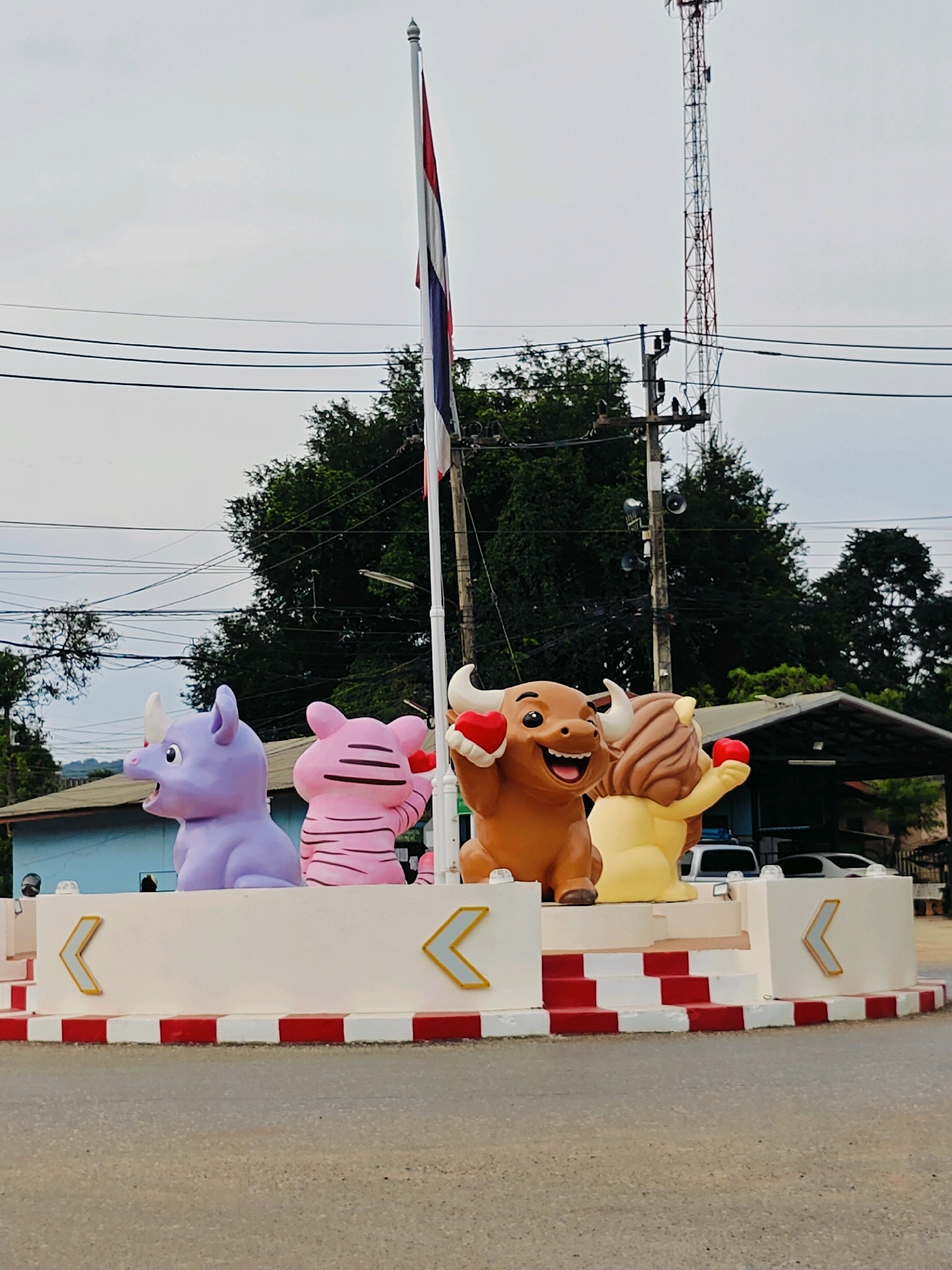 The image shows a roundabout in Tambon Mu Si, Thailand, featuring four cartoonish animal statues. From left to right, the statues are a purple rhinoceros, a pink pig, a brown bull, and a yellow lion. All statues are holding red heart shapes. The roundabout has a white border with red and white checkered stripes along its base. A flag pole with a Thai flag is visible behind the statues. Electrical wires and a communication tower are present in the background, along with trees and buildings. The scene appears to be outdoors during the daytime under an overcast sky.
