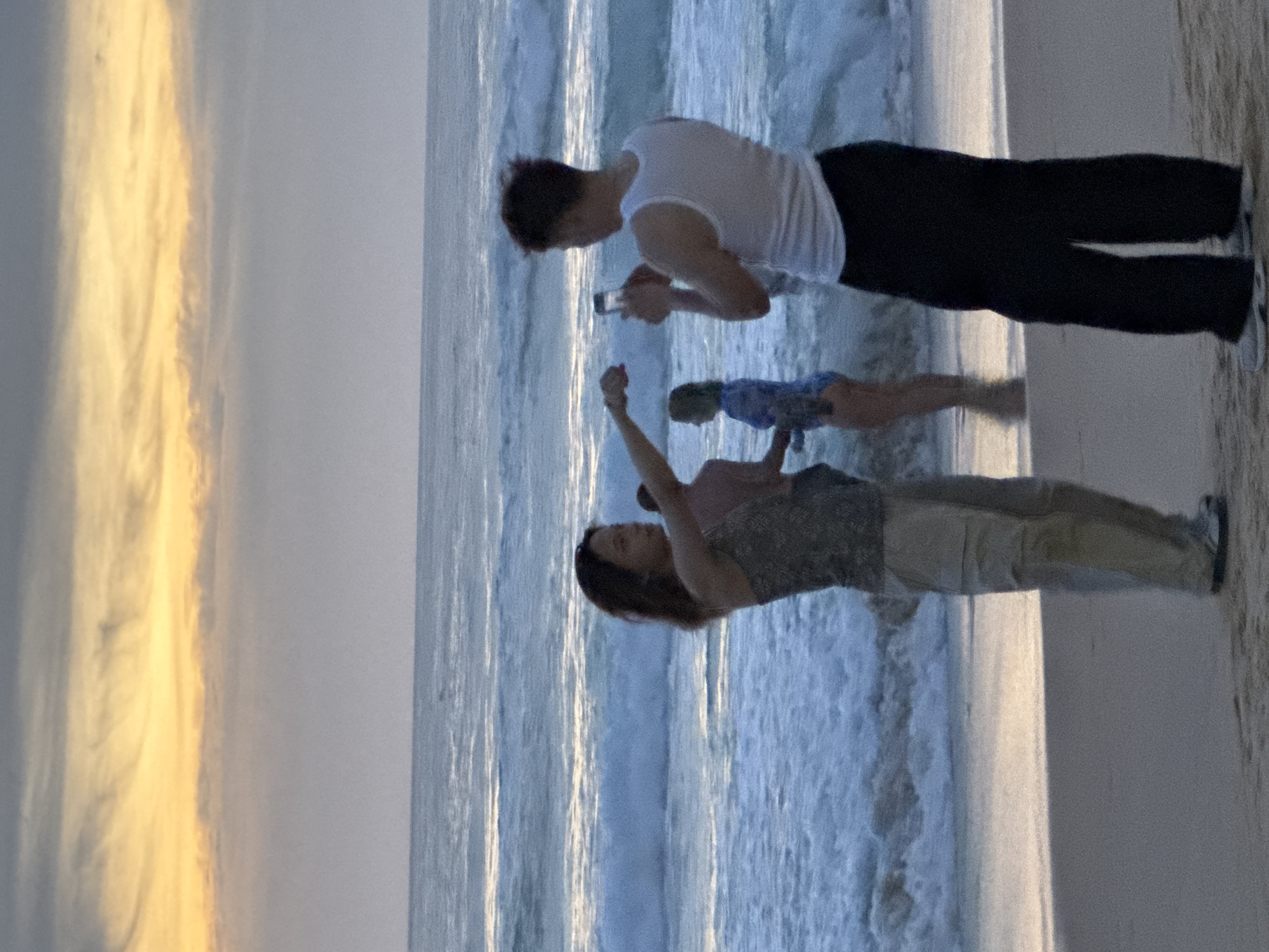 At sunset on Karon Beach, Thailand, a group of people are gathered on the sand near the water's edge. In the foreground, a woman in a patterned tank top and beige pants stands with her arm extended, holding a mobile phone, seemingly taking a photo or video. To her right, a man in a white tank top and black pants is also holding a mobile phone, appearing to take a picture of the woman. In the background, another person in a blue swimsuit is visible, and further out to sea, waves crash onto the shore under a sky painted with hues of orange, yellow, and gray. The lighting suggests it is late evening.