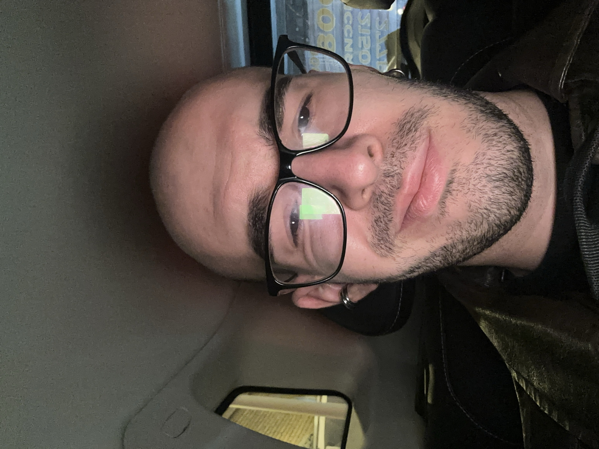 A close-up, angled selfie shows a light-skinned, bald man wearing black-rimmed glasses. He has a neatly trimmed, dark beard. He is likely in a vehicle, judging by the interior surfaces visible. A small section of a rearview mirror is visible in the lower part of the frame. The man is wearing a dark jacket or coat. The location in Moscow, Russia, cannot be determined from the visual information provided.