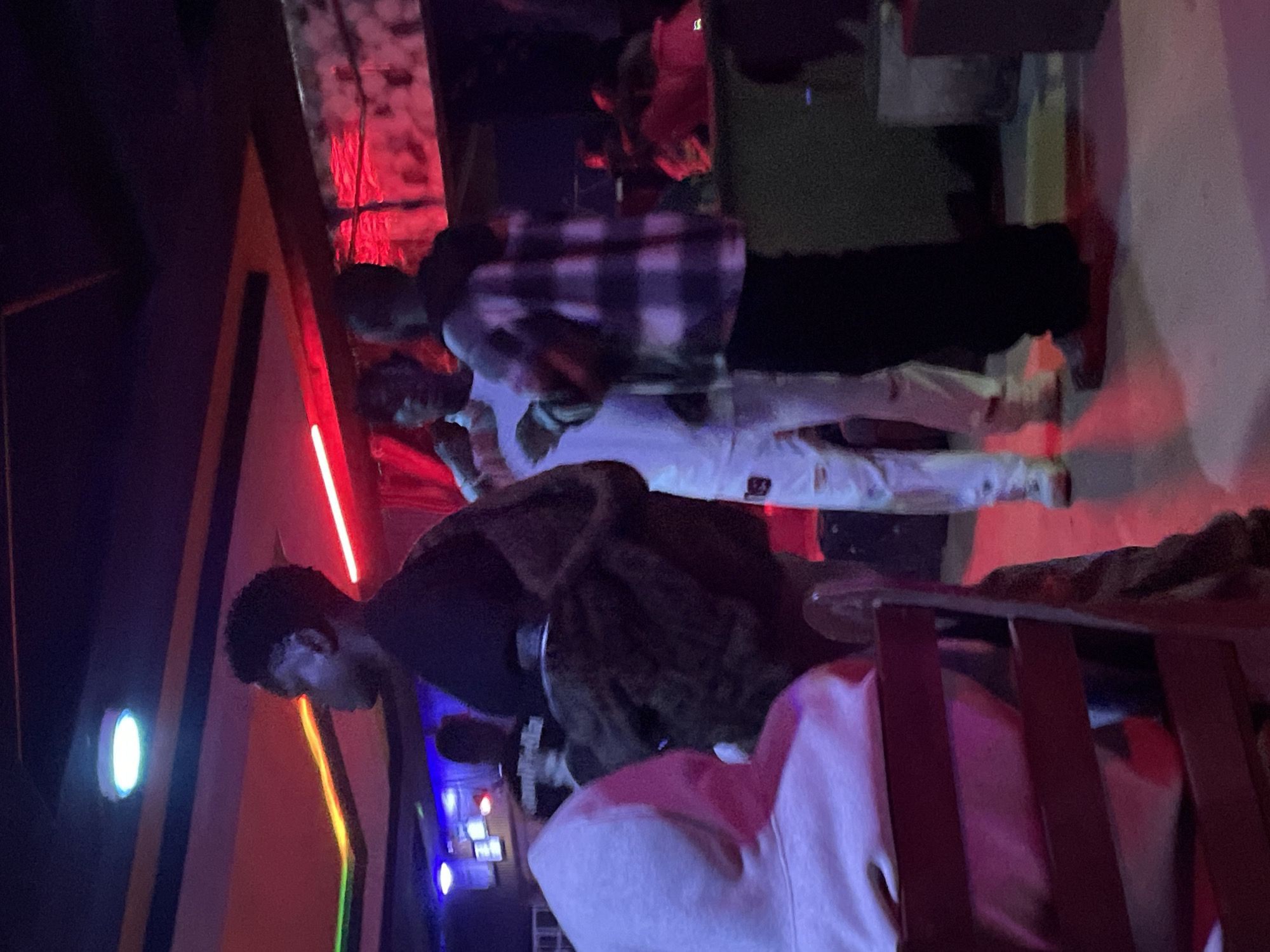 In Durumi, Nigeria, at a dimly lit establishment possibly a club or lounge, several individuals are present. The lighting is predominantly red and blue, casting a vibrant glow on the scene. In the foreground, a person wearing a light pink hoodie is seated, their back to the camera, facing a dark wooden chair with a pink cushion. To their right, a person in a dark jacket and what appears to be a furry lining is partially visible. Further back, two men stand in conversation. One, wearing a white t-shirt and white pants, has his left hand raised, possibly gesturing. The other man, wearing a checkered shirt and black pants, stands with his arms crossed. The environment includes industrial-style elements with exposed pipes and textured walls in the background. The floor is a concrete surface with distinct red and blue light reflections. The overall atmosphere suggests a casual social gathering or nightlife scene.