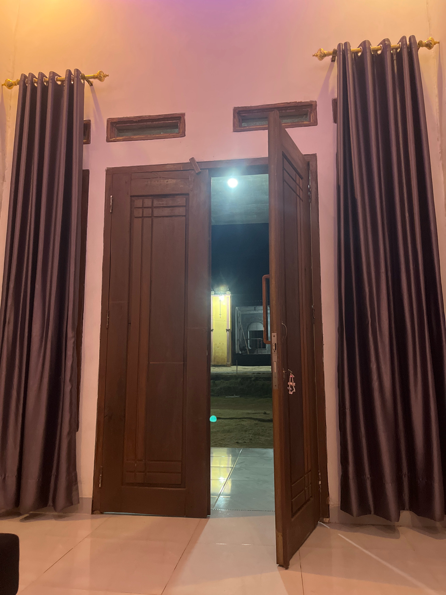 The image shows an open wooden double door leading outside from an indoor setting. The interior features light-colored tiled flooring and a pale pink wall. To the left and right of the door are dark brown curtains hanging from gold curtain rods. Above the door, on the wall, are two rectangular vents. The open doorway reveals a dimly lit outdoor area with a bright light source in the distance. The flooring transitions from tiles inside to a rougher surface outside. The overall atmosphere suggests an evening or night scene. The image is tagged with "Kebumen, Indonesia," implying the location is in that city. There are no people visible in the image, and no actions or interactions are depicted. The scene is static and observational.