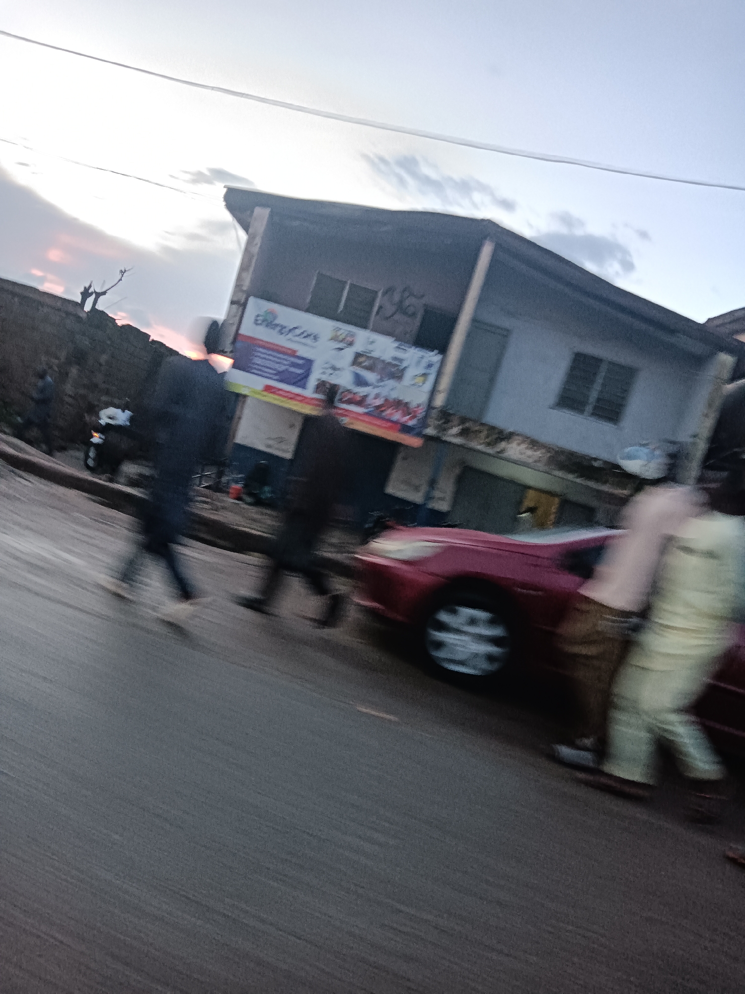 The image depicts a street scene in Jos, Nigeria, likely during dusk or dawn due to the soft, diffused lighting and the muted colors of the sky. The foreground is dominated by a wet asphalt road, suggesting recent rain. A blurred red car is visible in the middle ground, with its headlights off and the driver's side door ajar. Two individuals, also blurred due to motion, are walking on the road beside the car. One person, wearing a dark outfit, is walking away from the camera, while another, in a lighter-colored outfit, is walking towards the camera.
In the background, a two-story building stands on the right side of the street. The building appears to be a commercial establishment, with a sign that is partially obscured but appears to have colorful graphics and text, possibly advertising. The windows on the upper floor are visible, and the building has a weathered appearance. To the left of the building, a rough, unpaved area is visible, bordered by a low wall. A leafless tree is silhouetted against the sky in this area. The overall impression is one of a bustling urban environment with signs of daily life.