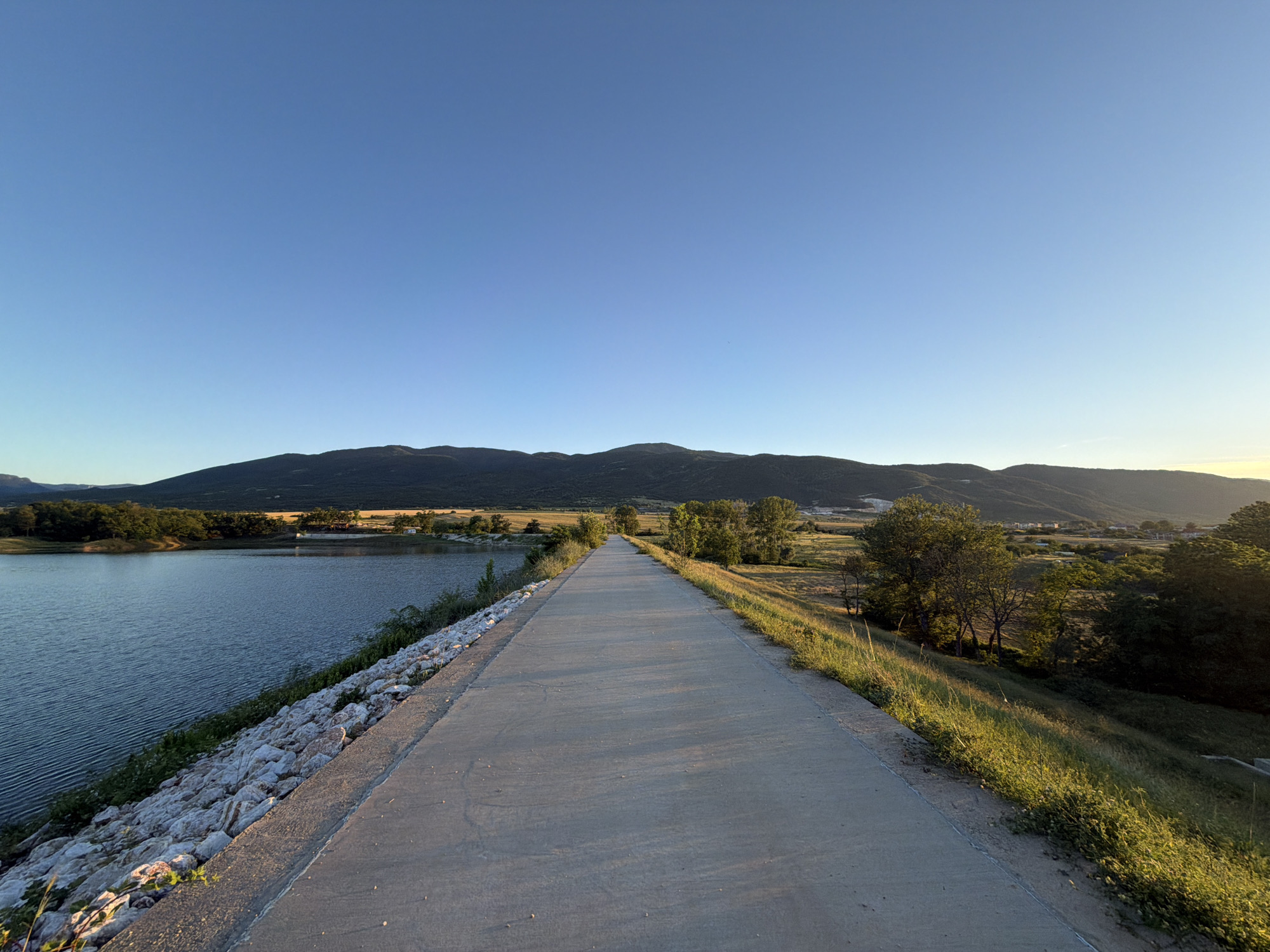 The image depicts a paved pathway running along the edge of a body of water, likely a reservoir or lake, with a mountainous landscape in the background. The pathway is made of concrete and appears to be a levee or dam. The water on the left is calm with visible ripples. The right side of the pathway is bordered by a grassy slope with scattered trees. In the distance, rolling hills covered in vegetation and some scattered buildings suggest a rural or semi-urban setting. The sky is clear and blue, indicating fair weather. The lighting suggests it is late afternoon or early evening, with the sun casting long shadows. This scene is located in the vicinity of Muldava, Bulgaria, a location known for its natural beauty and proximity to mountainous terrain.