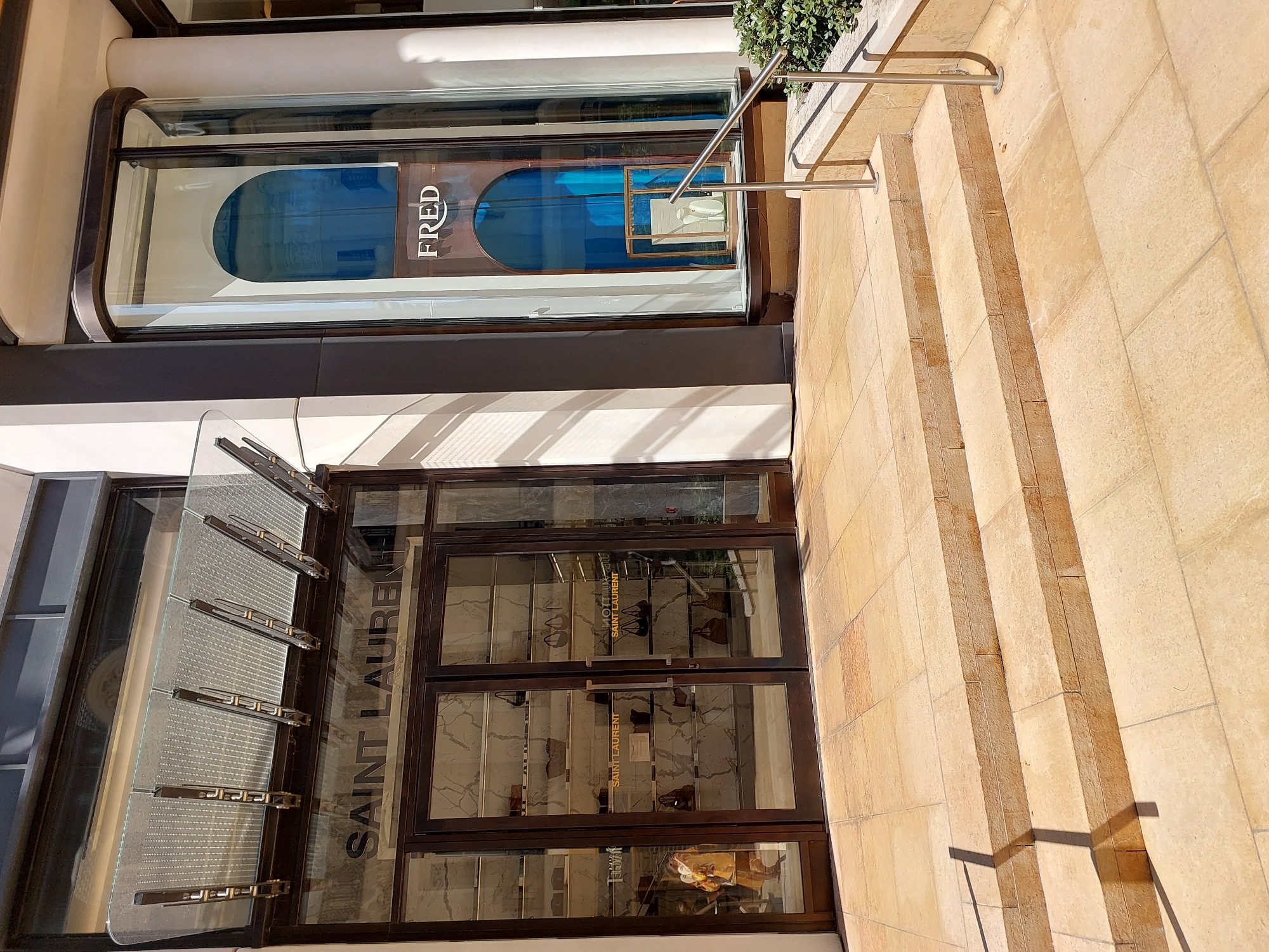 The image shows the exterior of two adjacent luxury retail stores, Saint Laurent and Fred, located in Monaco. The Saint Laurent store features a large glass storefront with double doors, displaying handbags on shelves. A person in a yellow outfit is visible inside the store. The Fred store has a curved glass display window with the brand name "FRED" prominently displayed. The stores are situated on a paved walkway with stone steps leading up to the entrance of Saint Laurent. A metal railing is present next to the steps. Green shrubs are visible to the right of the Fred store. The scene is illuminated by natural daylight.