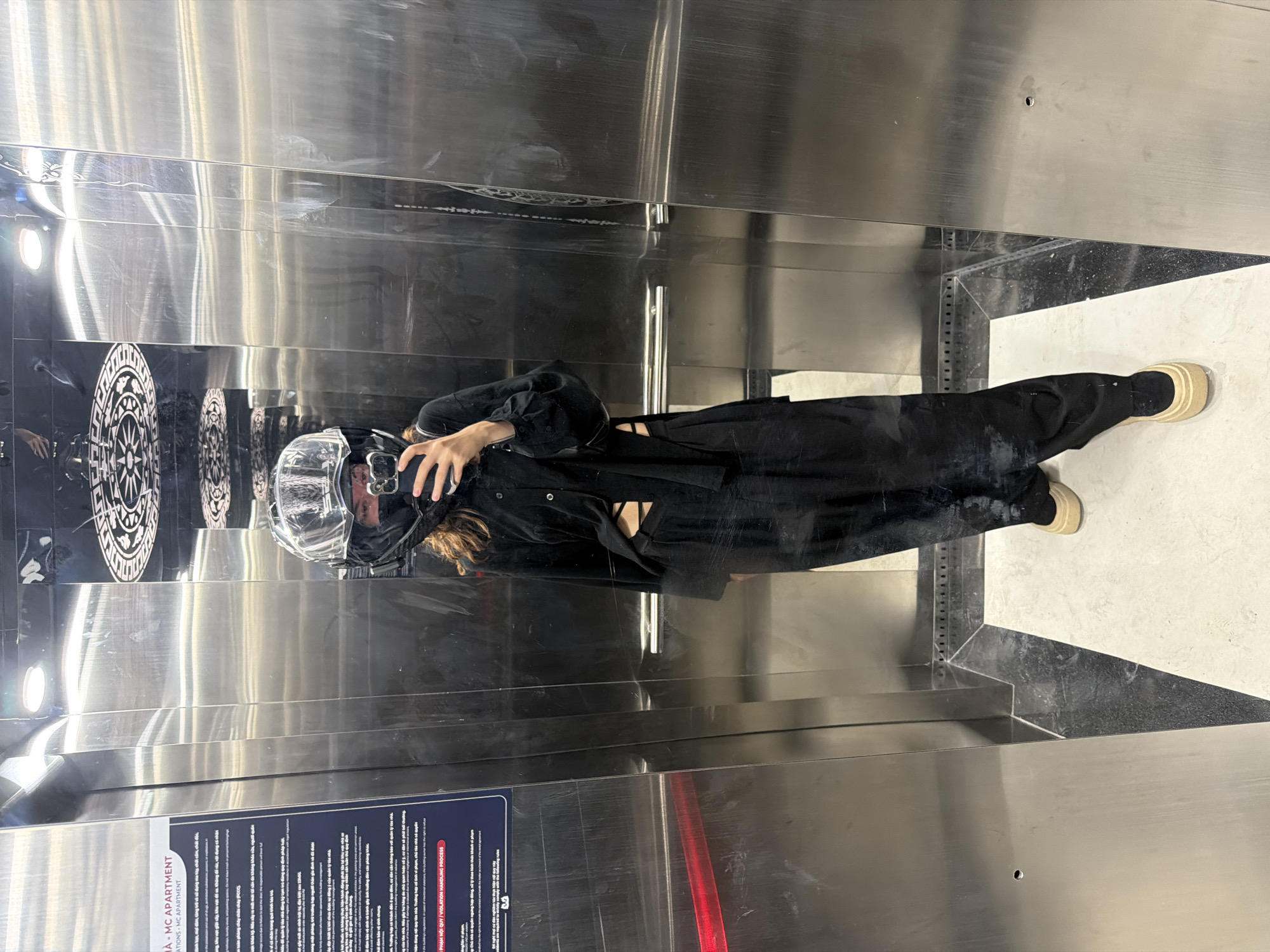 The image shows a person taking a selfie in what appears to be an elevator with stainless steel interior, based on the reflections and finishes. The person is wearing a black outfit that appears to be a jumpsuit and shoes with light-colored soles. The person is also wearing a motorcycle helmet, and holding a phone. The floor of the elevator appears to have marble floor trim along the bottom of the stainless steel wall. Additionally, some signs in Vietnamese are visible on the elevator wall. Based on the text in the signs, the photograph was taken in Đà Nẵng, Vietnam.