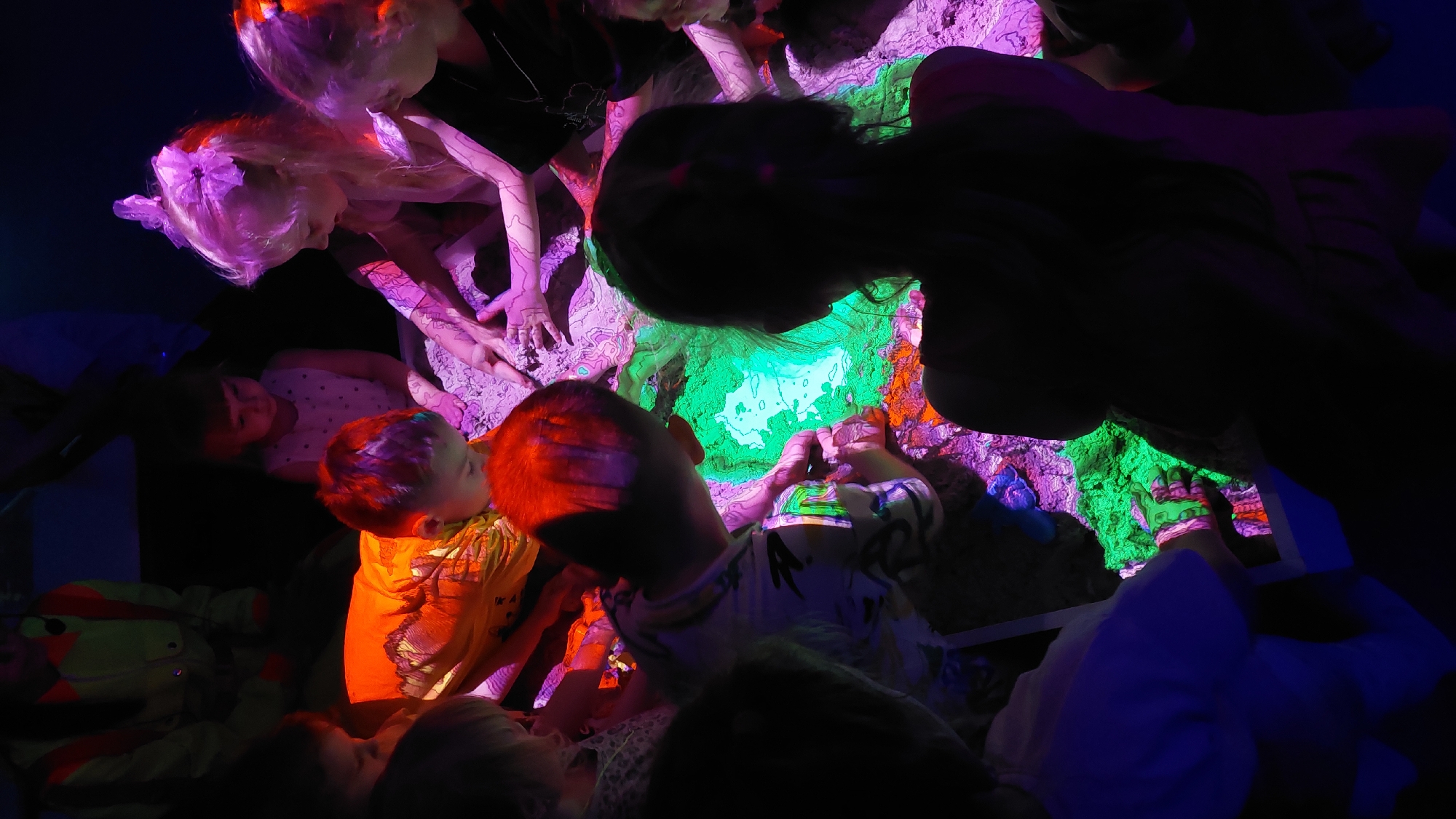This image depicts a group of children gathered around a brightly lit, sand-filled table or interactive exhibit, likely in Komsomolsk-on-Amur, Russia. The exhibit glows with vibrant green and purple light, illuminating the children's faces and hands as they interact with the sand. The children, varying in age, are leaning in, touching, and exploring the textured surface, their expressions suggesting curiosity and engagement. The surrounding environment is dark, accentuating the illuminated exhibit and the children's involvement. The overall scene suggests an educational or recreational activity designed to stimulate sensory exploration and imaginative play.