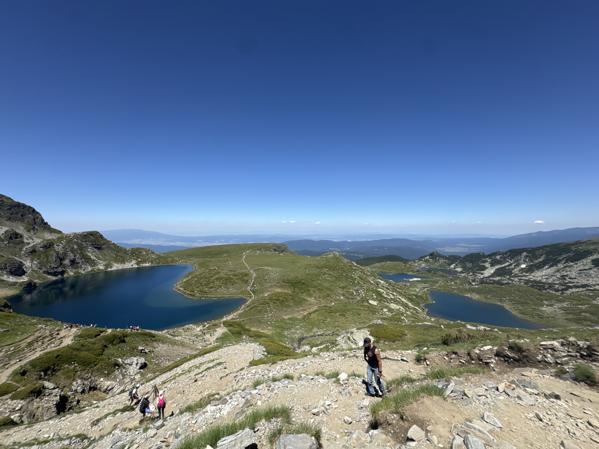 The image depicts a panoramic view of the Seven Rila Lakes in Panichishte, Bulgaria, under a clear blue sky. In the foreground, a rocky, uneven mountain trail is visible with several hikers making their way up and down. A person with a black t-shirt, black cap, and jeans is walking down the trail on the right side of the frame, holding a water bottle and a smartphone. Further down the trail, three individuals, two with backpacks and one in a pink backpack, are ascending. The middle ground showcases the stunning blue lakes nestled within lush green, rolling hills. The largest lake, known as the "Eye," dominates the left side of the image, reflecting the sky. Other lakes are visible further in the distance, surrounded by rocky slopes and vegetation. The background reveals a vast expanse of mountains and valleys, with a hazy horizon. The overall scene conveys a sense of natural beauty and outdoor recreation.