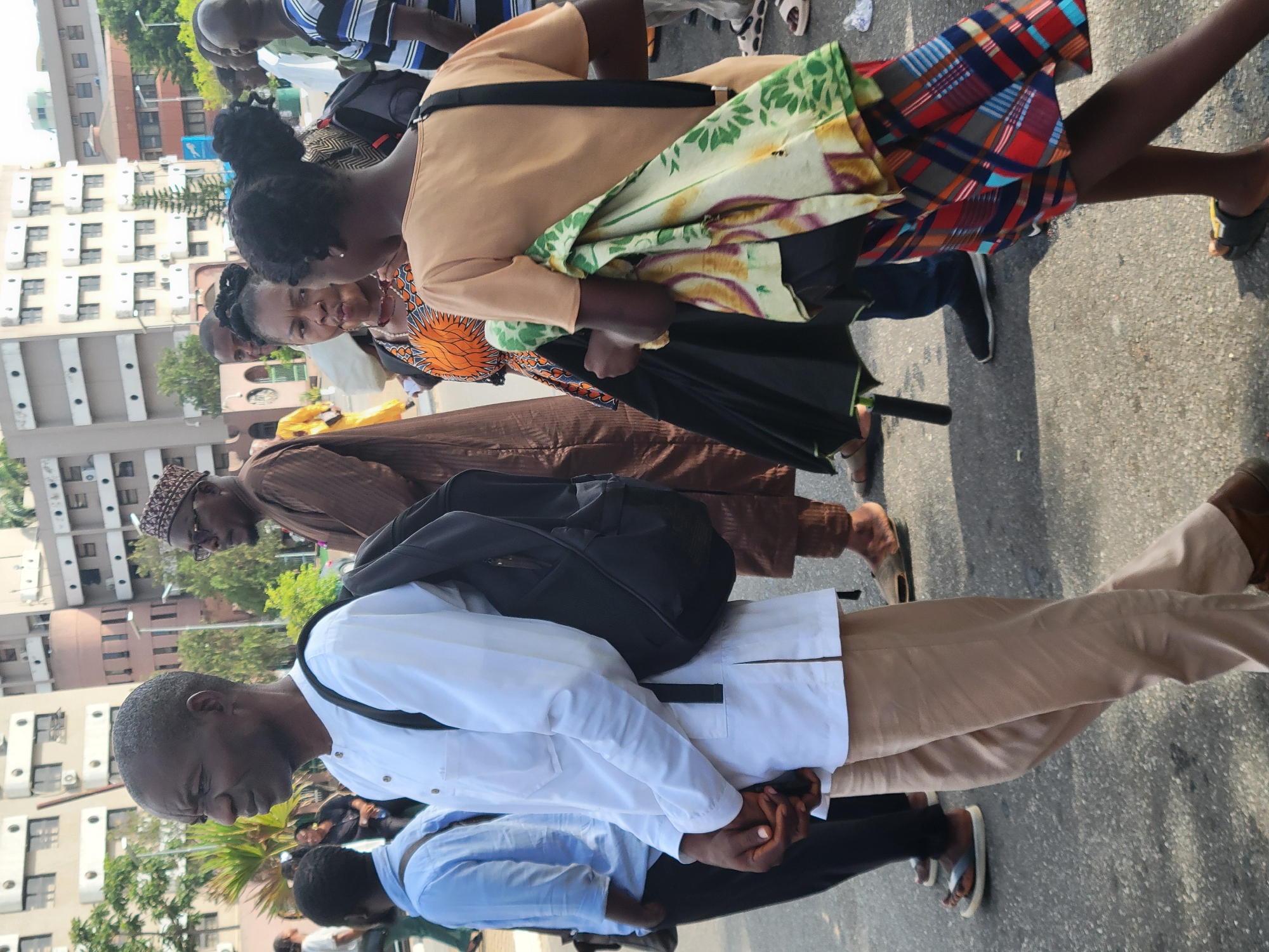 A medium shot of a group of people, mostly African, walking along a street in Abuja, Nigeria.
In the foreground, a man in a white shirt and khaki pants with a black backpack walks with his hands clasped together. Behind him is a man in a brown robe with a patterned hat. A woman with a colorful dress and a green patterned scarf is visible behind the second man. More people can be seen in the background.
The street is paved and appears to be in an urban area, with tall buildings visible on the left side of the frame. The lighting suggests it is daytime.