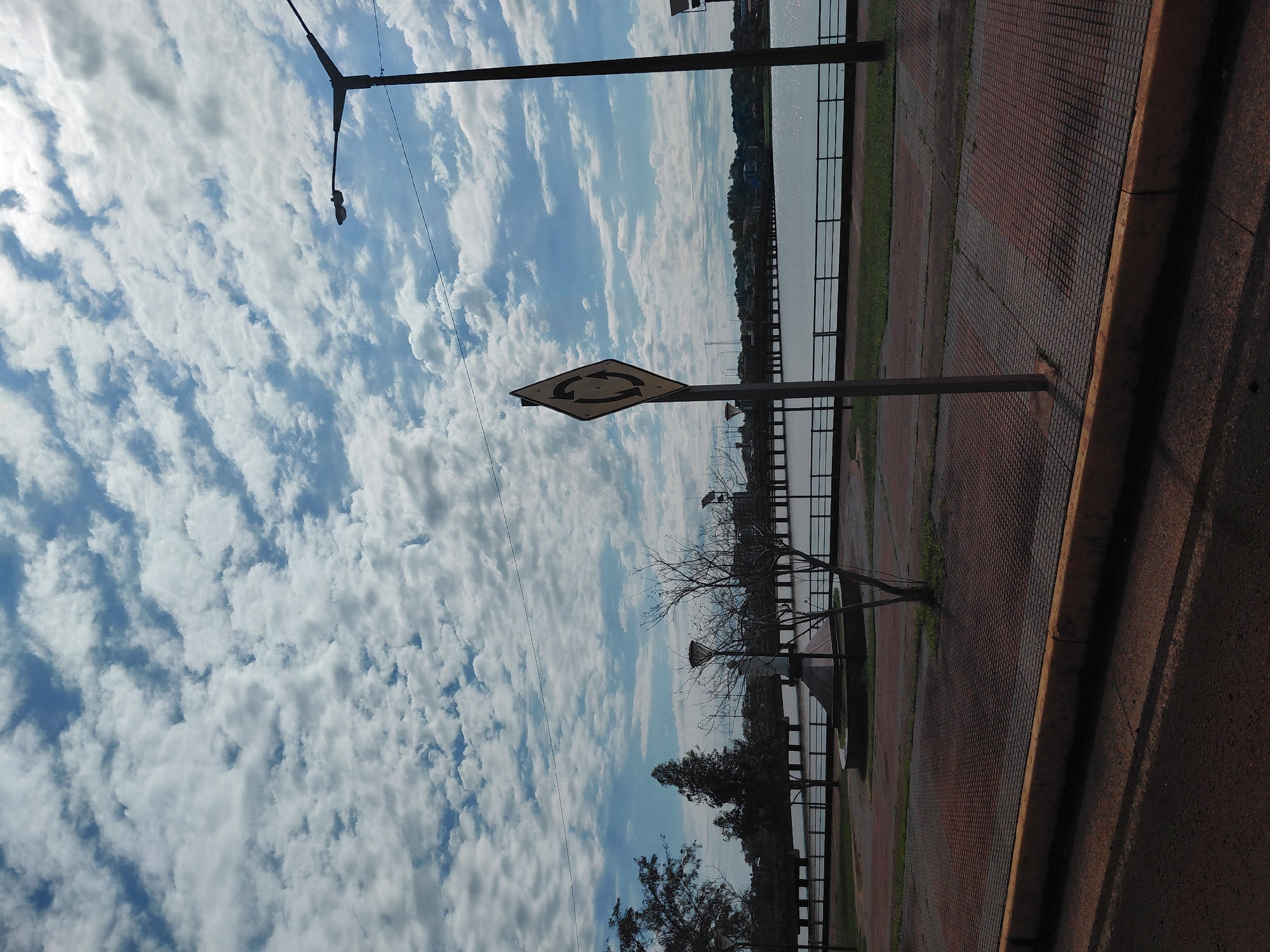 The image depicts a view of the Costanera de Encarnación, a waterfront promenade in Encarnación, Paraguay. The scene is characterized by a bright, cloudy sky with scattered white clouds against a blue backdrop. A prominent roundabout signpost stands in the foreground, indicating a traffic circle. To the right, a lamppost, a common fixture along the Costanera, is visible.
In the middle ground, a body of water, likely the Paraná River, stretches across the frame, bordered by a railing and a grassy area. Beyond the water, a bridge and a distant treeline suggest the urban landscape of Encarnación. The foreground features a paved walkway made of reddish-brown tiles, with a yellow curb separating it from a darker paved surface, possibly a road.
Several bare trees are scattered throughout the scene, some appearing pruned or naturally stunted. A monument, partially obscured by a tree, is visible near the water's edge. The overall atmosphere is serene, with no people or vehicles immediately apparent. The lighting suggests it is daytime, with shadows cast on the paved surfaces.