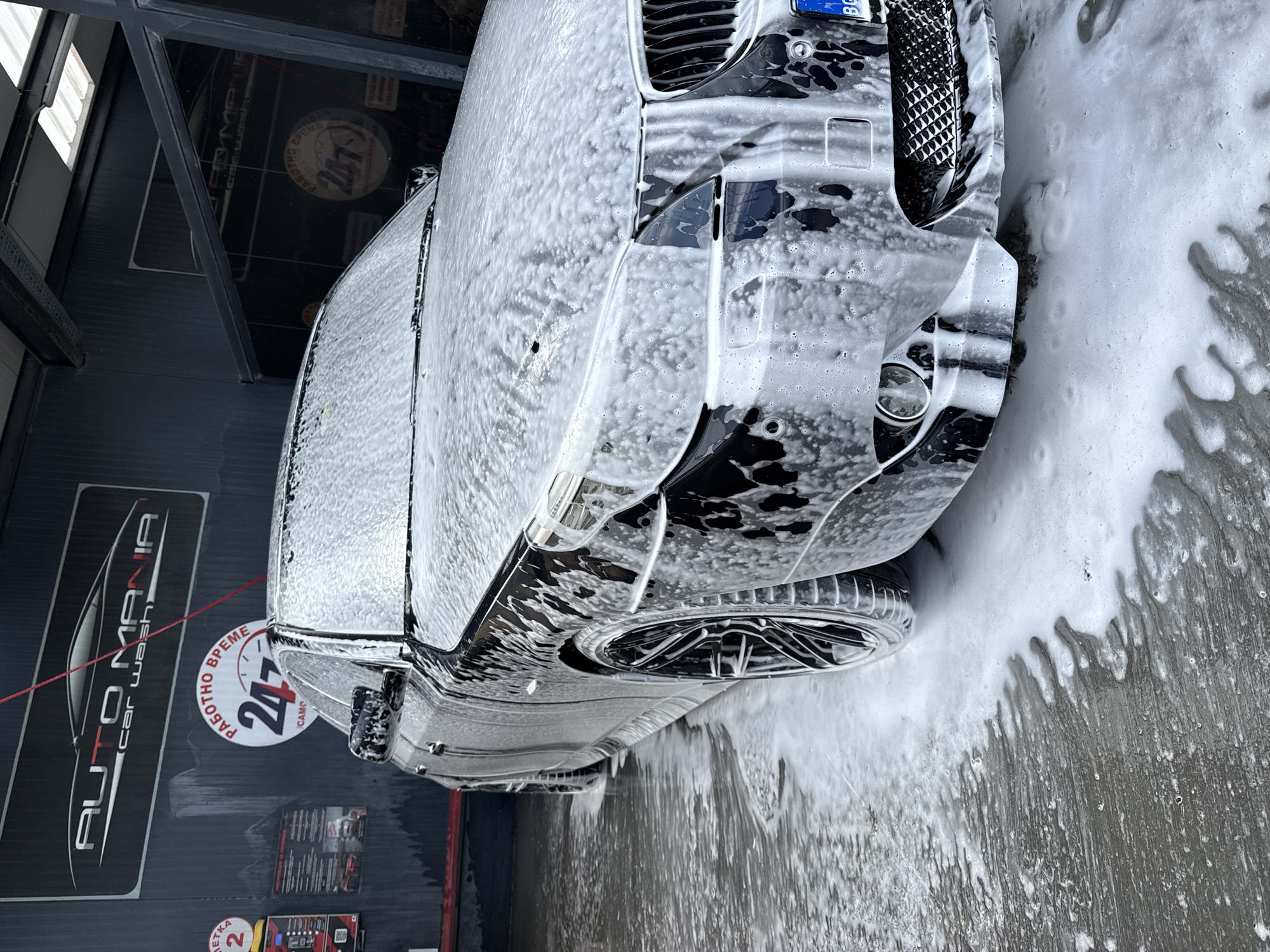 A black car is covered in white soap suds at the Auto Mania Car Wash in Gabrovo, Bulgaria. The car's front end, including the grill, headlights, and hood, is visible. The car is parked on a gray, textured surface also covered in soap suds. A sign for Auto Mania Car Wash, featuring a car design and the phrase "24/7" in Bulgarian, is visible in the background on a gray wall. Other signs and equipment are also seen on the wall.