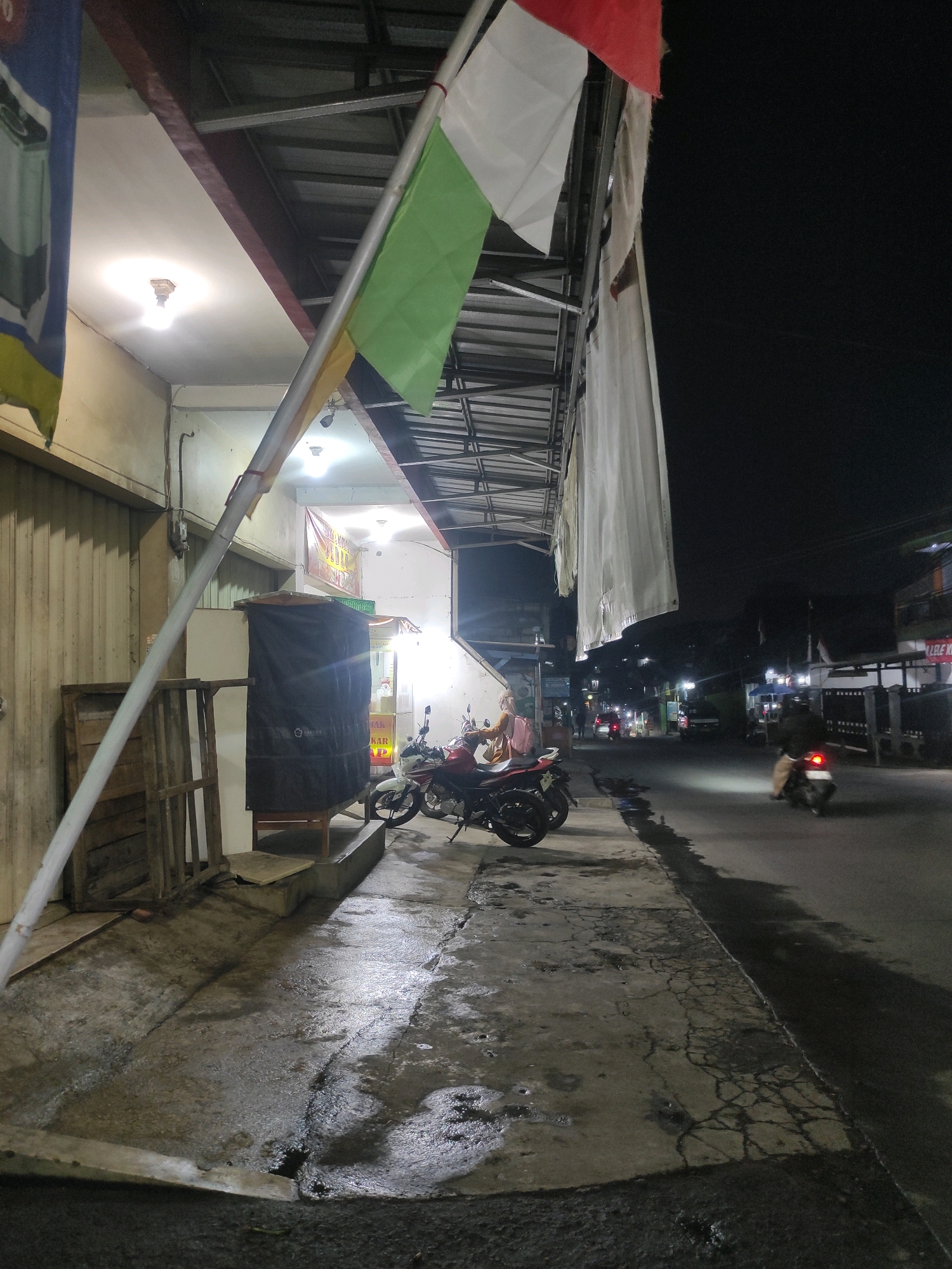 This is a night photograph taken in the city of Padalarang, Indonesia. The scene depicts a street with a sidewalk in front of a commercial building. Several motorcycles are parked on the sidewalk, and one person is sitting on a motorcycle. A flag is hanging from a pole in front of the building. The street is wet, indicating recent rain. Buildings with lights are visible in the background, and there are streetlights illuminating the road. The overall impression is of a quiet, residential or small commercial street at night.