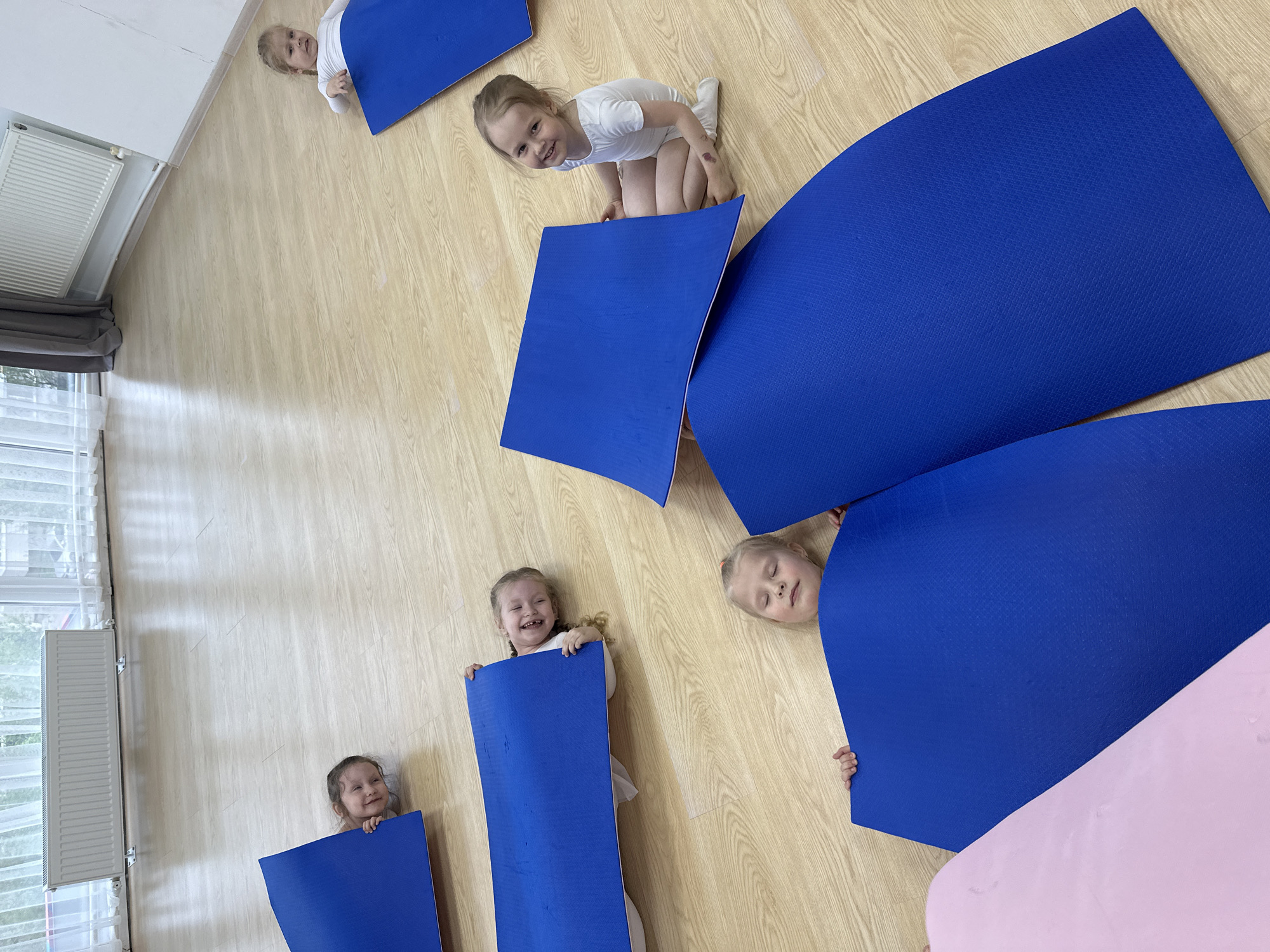 Five young girls are present in a brightly lit, indoor room in Votkinsk, Russia. The room has a light, wood-patterned floor and white walls. A window with closed blinds is visible in the lower left corner of the frame. The girls appear to be playing with blue and pink yoga mats. Each girl is partially obscured by a mat, either peeking over or holding it in front of them. Their expressions range from smiling and laughing to seemingly resting. One girl is kneeling on the floor.