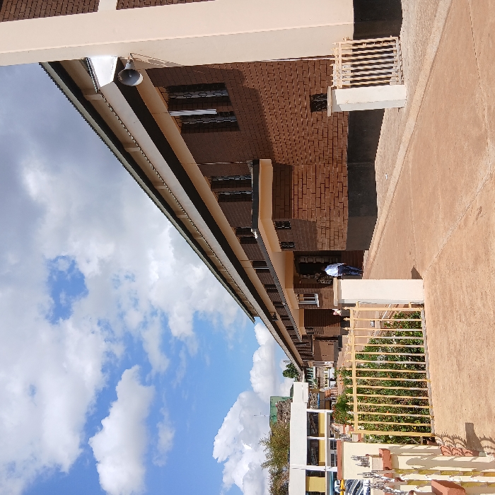 The image depicts an outdoor scene featuring buildings, sky, and ground, captured on a bright day. The primary subject is a multi-story building with reddish-brown brick walls and light beige or off-white trim, extending horizontally across the frame from left to right. This building features numerous dark-paned windows, some appearing to have internal blinds or coverings. A continuous, light-colored gutter or overhang system runs along the roofline on the left side of the building. A dark, dome-shaped security camera is mounted under this overhang towards the left.
In the foreground and to the right, there is a wide expanse of light brown or reddish-brown paved ground. Another section of a building, similar in construction to the main one, is visible on the right, featuring a small, light-colored railing or balcony. One person is standing near this right-hand building, wearing a dark top and light pants, appearing to look down or away from the camera. Further to the right and in the lower foreground, a prominent yellow metal gate with vertical bars and some decorative elements is visible, behind which green foliage and bushes can be seen, indicating landscaped areas.
A significant portion of the upper part of the image is filled with a bright blue sky dotted with fluffy white cumulus clouds, suggesting clear and sunny weather. The overall lighting indicates it is daytime, likely mid-morning or afternoon. No discernible text is visible in the image. The scene conveys a calm, everyday outdoor environment.