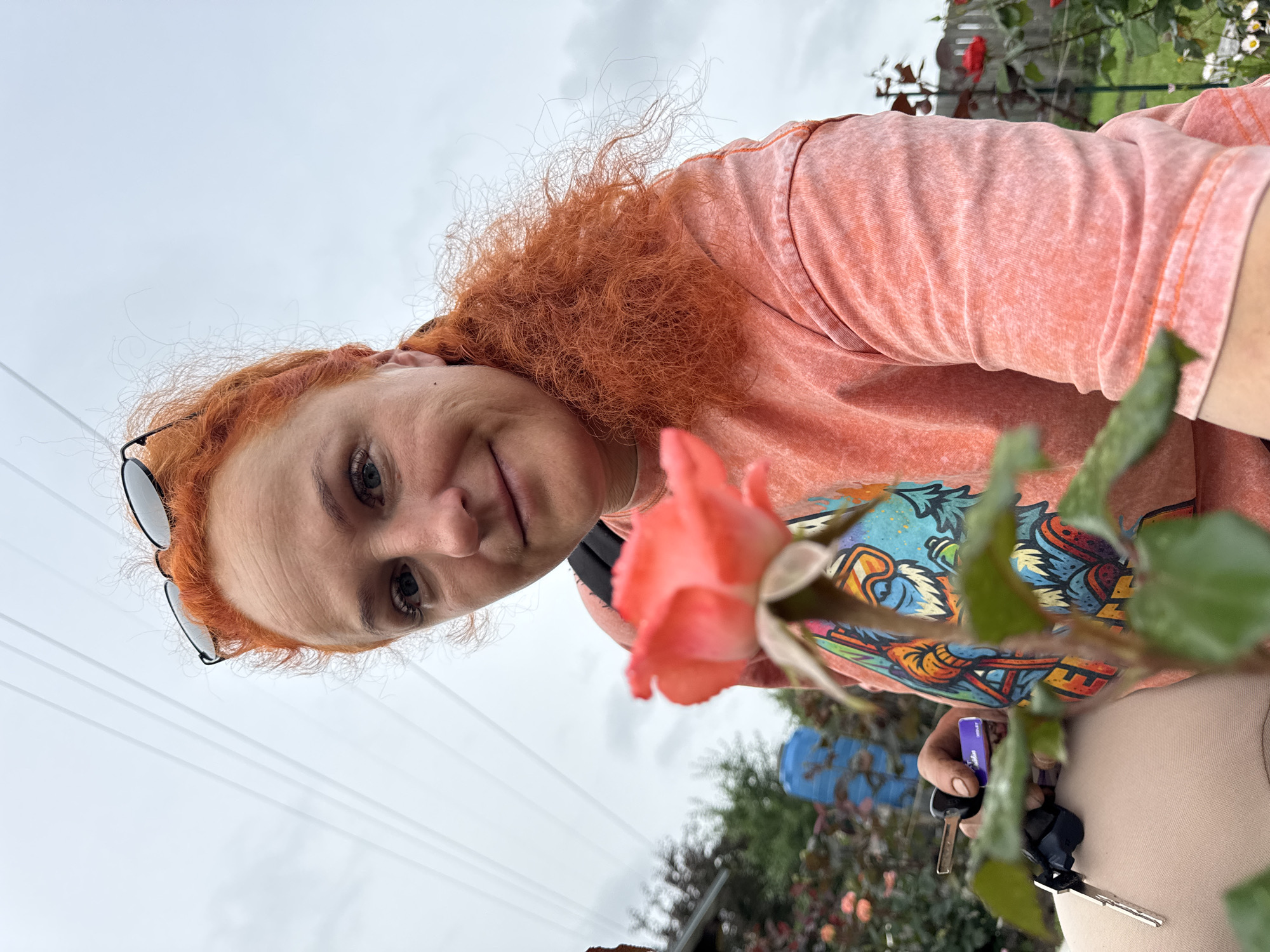 A woman with vibrant orange, curly hair and fair skin is captured in a close-up, low-angle shot. She is wearing a faded coral-colored t-shirt with a graphic print of a cartoon character wearing goggles and a ski outfit. Sunglasses are perched on her head, and she is smiling slightly at the camera. In her left hand, she holds a set of keys, one of which has a purple tag. In the foreground, a delicate pink rose is in sharp focus, its petals gently unfurling. Behind the woman, a blurred background reveals a garden with more roses, a blue barrel or tank, a house with a red-tiled roof, and a wooden fence. Overhead, a network of power lines stretches across an overcast sky. The setting appears to be a residential area, likely a garden or yard, with no specific identifying landmarks for Chaikovsky, Russia, visible in the image.
