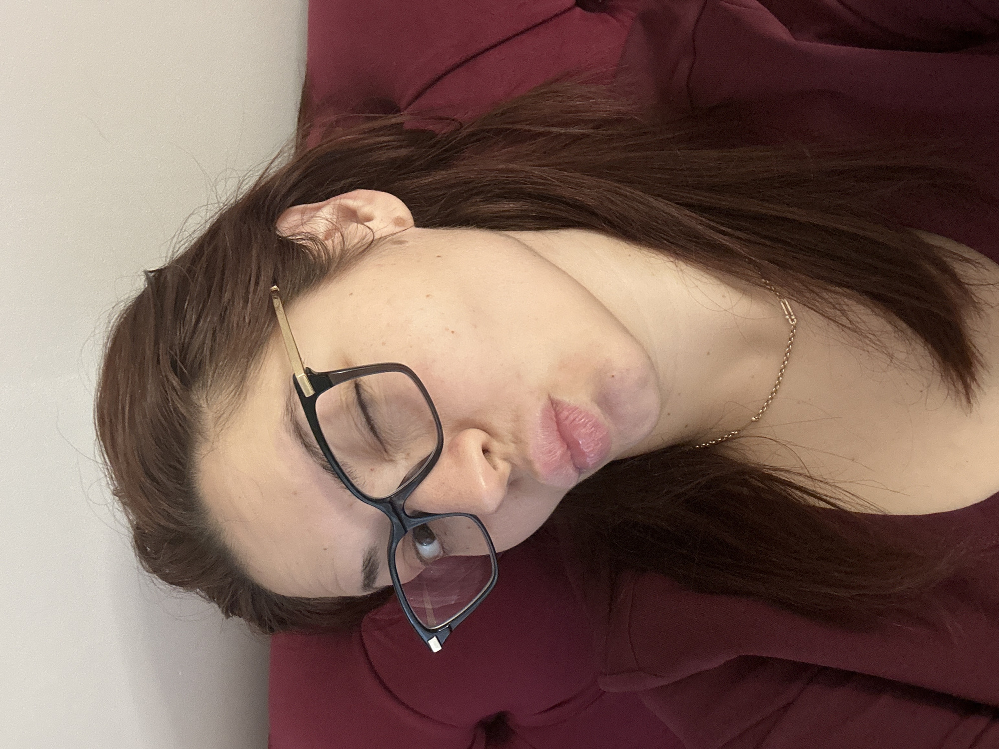 A woman with auburn hair and wearing glasses lies on a red surface. She is wearing a gold chain necklace. The background is a plain white wall. There is no evidence indicating the location is Sofia, Bulgaria.