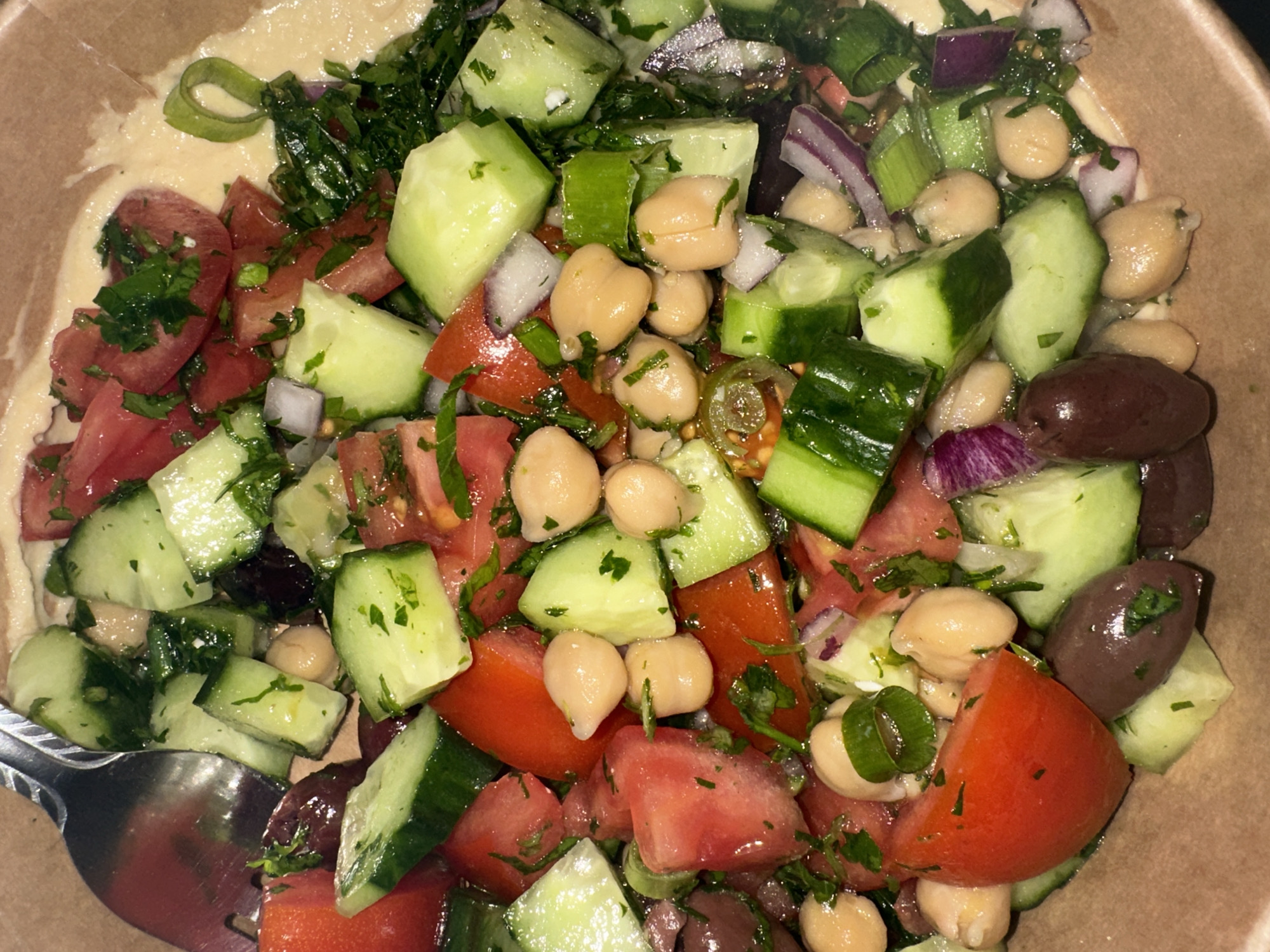 A close-up, overhead shot depicts a salad served in a tan bowl with hummus along the edge, likely in Sofia, Bulgaria. The salad consists of diced tomatoes, cucumbers, red onion, chickpeas, sliced olives, and green herbs, possibly parsley and scallions. A silver spoon is visible in the bottom left corner. The lighting suggests an indoor setting.