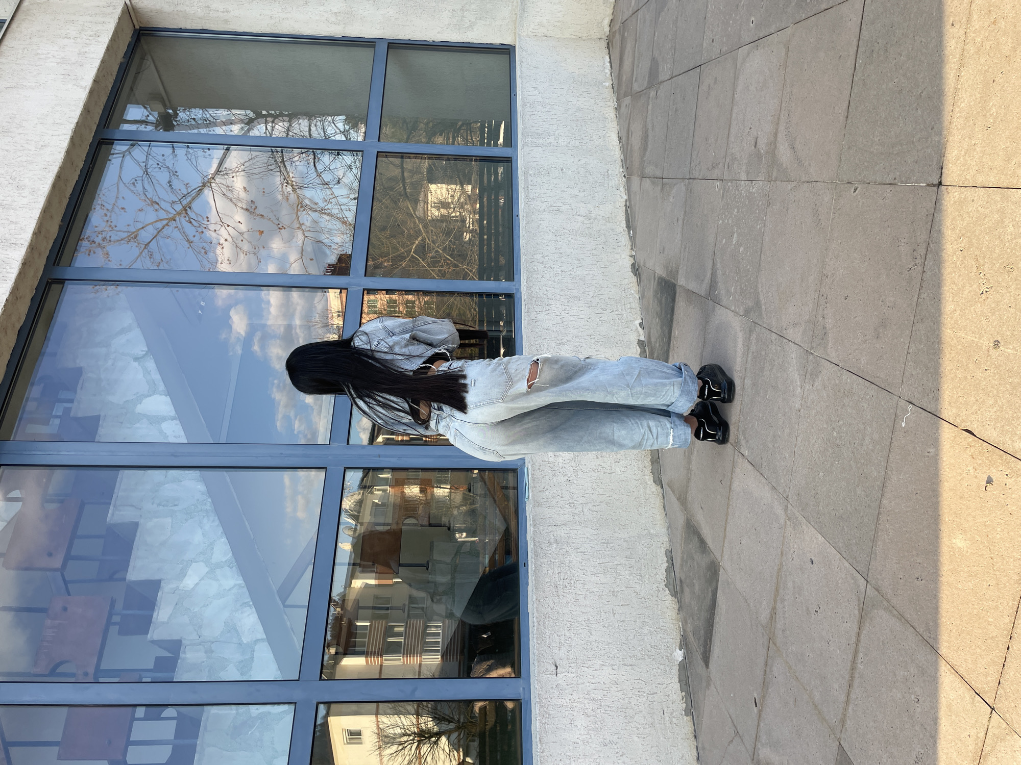 A woman stands outside a building with large paneled windows in Zlatograd, Bulgaria. She is facing the windows, her back to the viewer. She has long, dark hair and wears a denim jacket and ripped jeans with black shoes. The building windows reflect the outside environment, including buildings and trees. The woman is standing on a tiled sidewalk, which is partially in shadow.