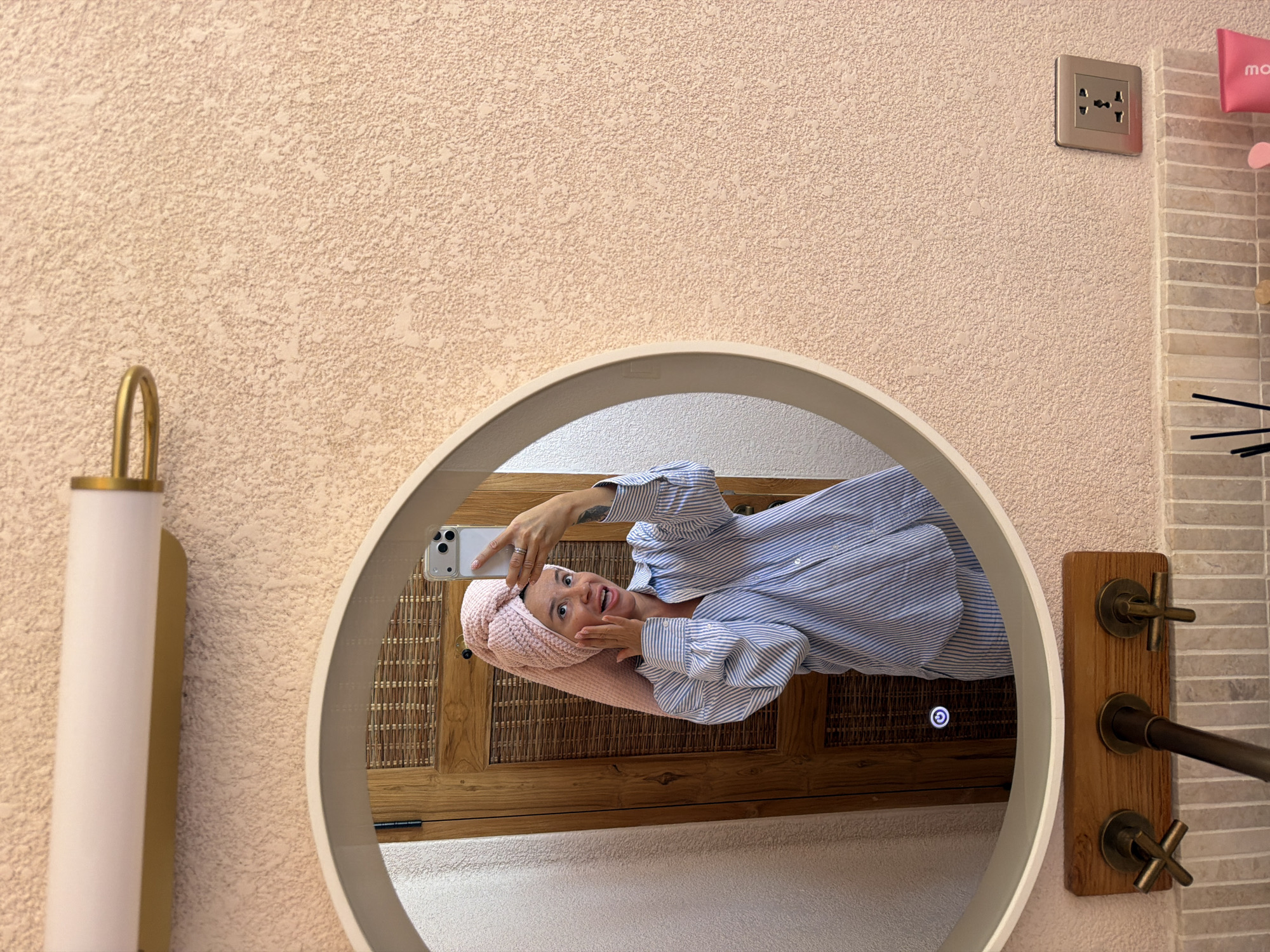A young woman with a pink towel wrapped around her head and a blue and white striped shirt is taking a mirror selfie in a bathroom. She is holding her phone in her right hand and her left hand is on her cheek. She is making a funny face, with her tongue sticking out and her eyes wide open. The bathroom has a pink textured wall, a round mirror, and a wooden cabinet. There is also a light fixture above the mirror and a power outlet on the wall. The floor is tiled with small, rectangular tiles. A wooden vanity with three faucets is visible at the bottom of the image. The overall scene suggests a casual and playful moment.