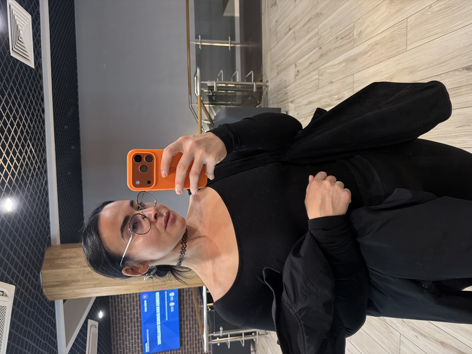 A woman is taking a selfie with an orange phone case. She is wearing glasses, a black choker, a black shirt, and a black jacket. The background is a room with grey walls, a wooden floor, and a black and white patterned wall. There is also a TV screen and a metal bicycle rack. The location may be a gym or recreational facility in Saint Petersburg, Russia.