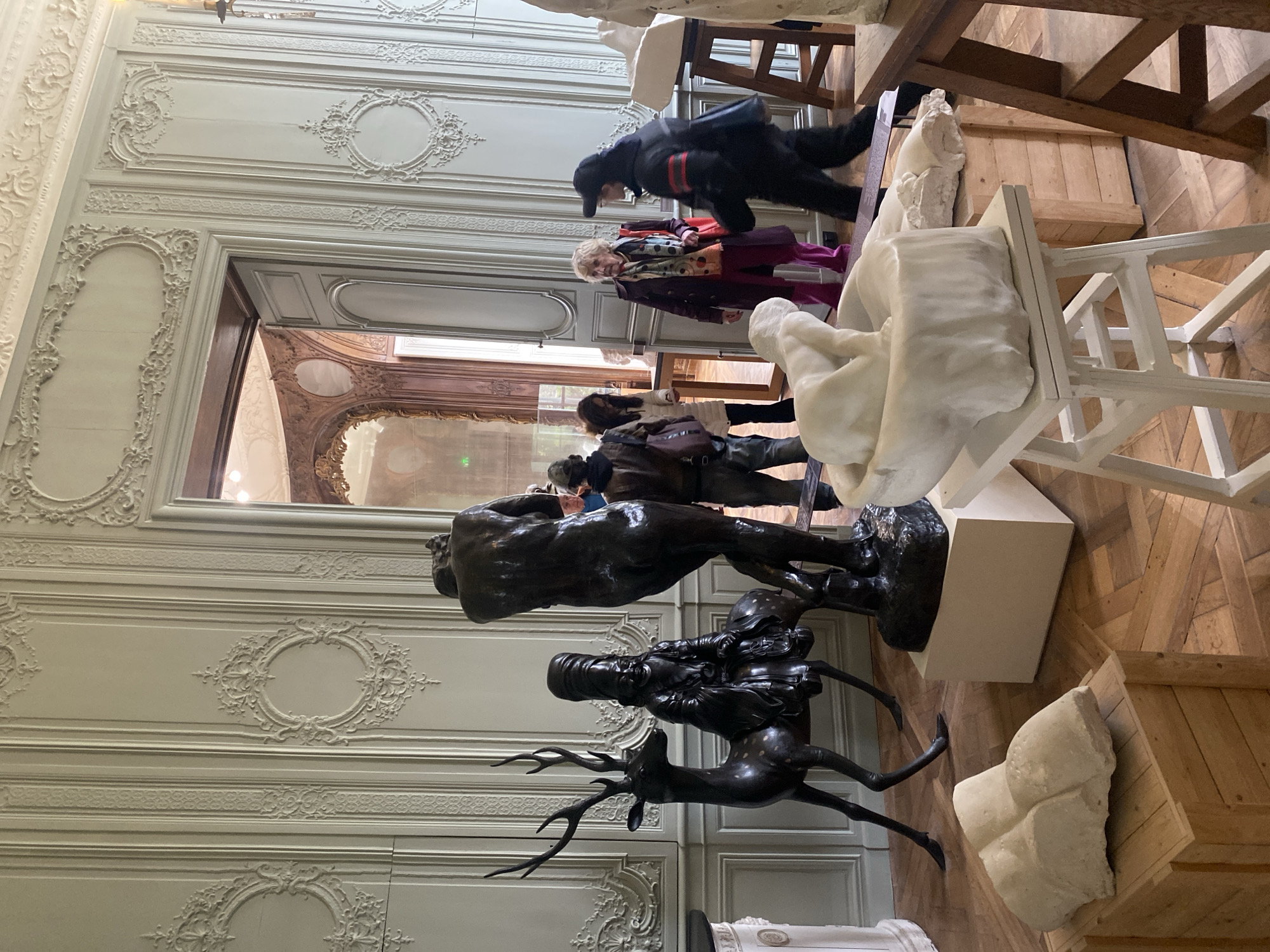 The scene depicts the interior of a museum gallery, specifically the Musée Rodin in Paris, France. The room features high ceilings and pale grey-green walls adorned with elaborate classical French boiseries, including decorative moldings and empty oval frames. A large arched doorway to the right reveals an adjacent room with darker, ornate wood paneling and a large mirror reflecting interior light. The floor is made of light-colored parquet wood.
Several sculptures are displayed throughout the room. In the mid-ground, a large, dark bronze sculpture of a standing deer with prominent antlers faces left, carrying a smaller, dark bronze figure with elongated headwear on its back. To its right, a large, dark bronze nude male figure, seen from the back, stands on a light pedestal; this figure is muscular and appears to have its hands behind its head. In the foreground, a rough-hewn white marble or plaster sculpture, possibly a torso, rests on a wooden crate. To the right, a white marble sculpture of a reclining figure is displayed on a white wooden stool, with another white marble fragment on an adjacent wooden crate. Additional white sculptures are partially visible on a wooden stand to the far right.
Five individuals are visible. In the background, two people are partially obscured near the doorway. In the mid-ground, an older woman with blonde hair, wearing a plum-colored coat, colorful scarf, and pink trousers, stands observing. To her right, another person wearing a black jacket with red stripes on the sleeve and a black cap walks towards the right, appearing in motion. The individuals are circulating, observing the sculptures within the museum setting.