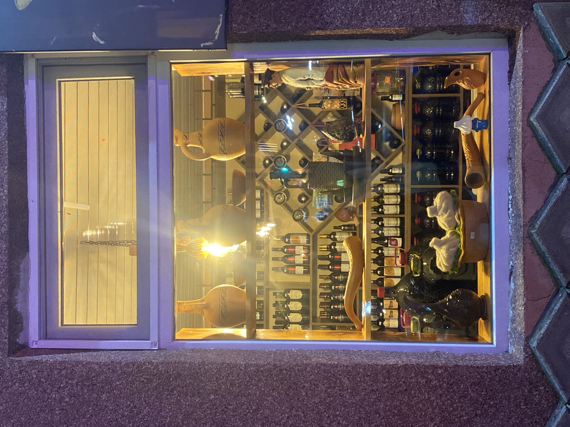 The image shows a shop window display in Batumi, Georgia, featuring a variety of items, primarily bottles and decorative objects. The window is illuminated from within, casting a warm glow on the display.
On the top shelf, there are several large ceramic jugs, two of which are prominently visible. One jug is a light brown color with dark blue decorative bands. Next to it, another jug is visible, and behind them, there appears to be a bowl. The background of this shelf has horizontal blinds.
The second shelf displays numerous wine bottles arranged in a wine rack. The bottles are dark in color, and some have visible labels. Above the wine rack, there are circular decorative elements. To the right of the wine rack, a figurine of a man in traditional clothing is standing. He is wearing a hat, a light-colored jacket, and a red and yellow patterned lower garment.
The third shelf also contains many wine bottles, similar to the second shelf, arranged in rows. In the center of this shelf, a long, curved, horn-shaped decorative object is placed. To the left of the horn, there are some dark glass bottles.
The bottom shelf displays a collection of dark glass bottles, some with colored caps. In the foreground of this shelf, there are two light-colored, bulb-shaped objects that resemble garlic or onions, placed in a small brown bowl. To the right of these objects, there is a small blue and white item and a small orange ceramic pitcher.
The shop window is framed by a textured, speckled facade. The overall impression is a display of wine, traditional crafts, and decorative items, likely intended to attract customers to a wine shop or a similar establishment. The lighting suggests it is nighttime or a dimly lit interior.