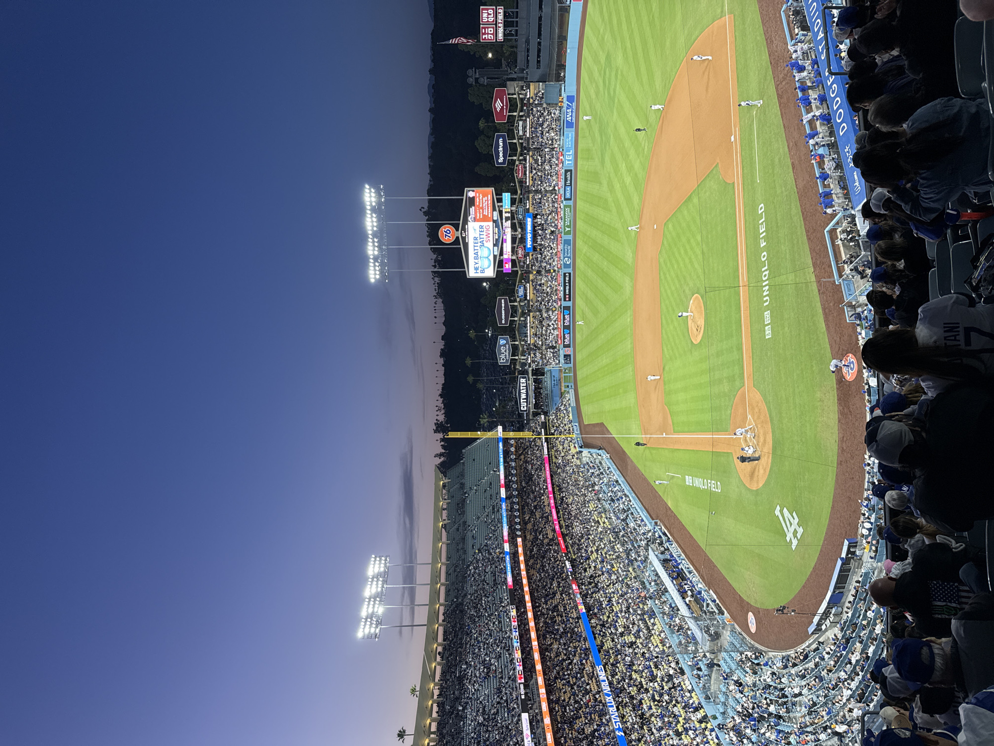 This photograph depicts an aerial view of a baseball game at Dodger Stadium in Los Angeles, United States. The stadium is illuminated by bright overhead lights, contrasting with the twilight sky. The baseball field is marked with "UNIQLO FIELD" and the "LA" logo. Spectators fill the stands, with many wearing blue attire, consistent with the Los Angeles Dodgers' team colors. Various advertisements and scoreboards are visible beyond the field, along with trees and a hillside in the background.