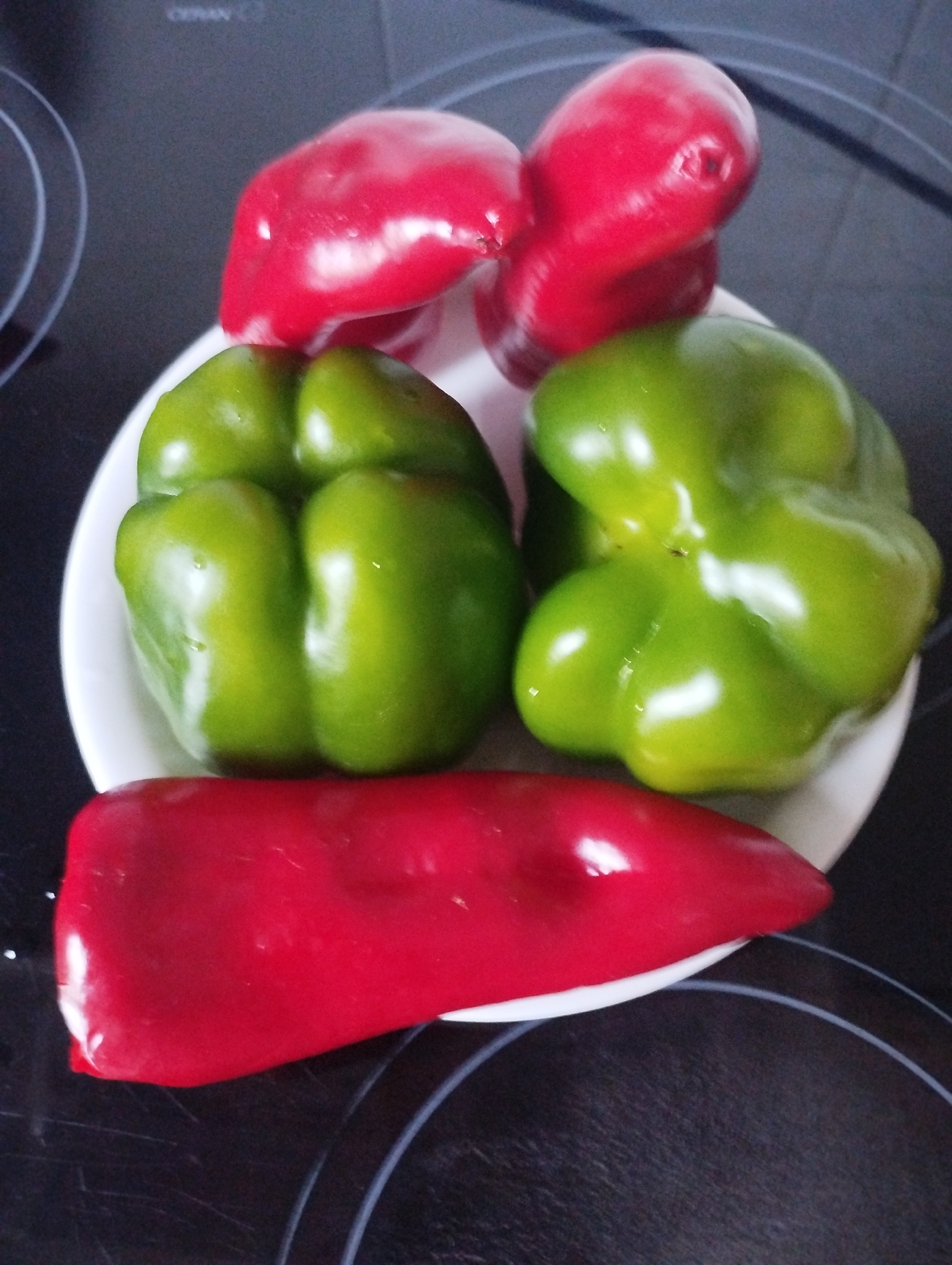 A high-angle shot shows a white plate holding five bell peppers on a black stovetop. Two bright red bell peppers are at the top of the plate, one slightly behind and to the left of the other, both appearing somewhat wrinkled. Below these, two vibrant green bell peppers are positioned side-by-side, their surfaces smooth and glossy. Finally, a single, elongated red bell pepper lies horizontally at the bottom of the plate, its shape distinct from the bell peppers above. The black stovetop has visible circular heating elements. The scene is static and appears to be a still life arrangement of produce. No people are present, and no specific location in Biasca, Switzerland can be identified from the image.