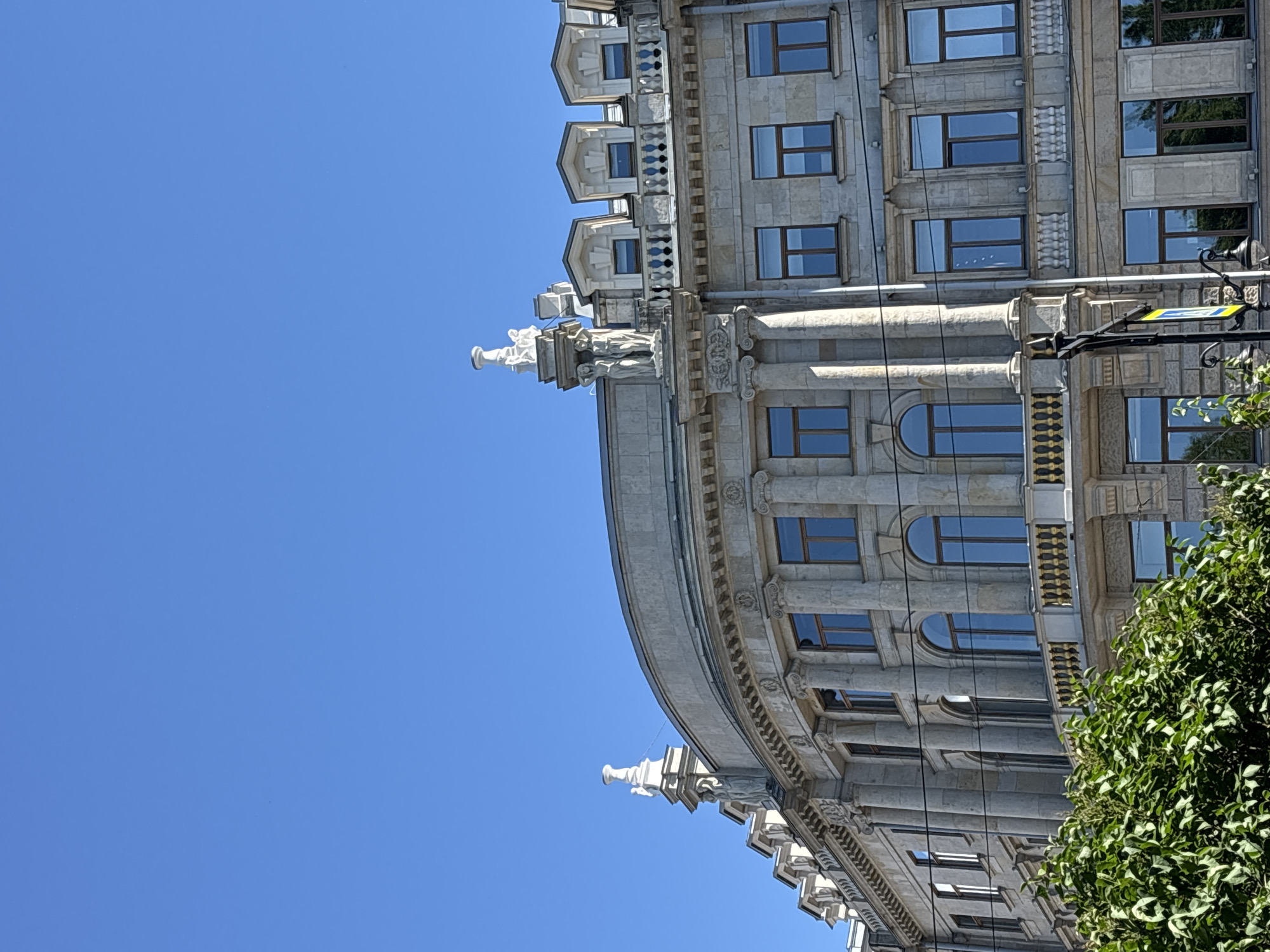The image shows a close-up view of the facade of a grand, historic building in Saint Petersburg, Russia. The building is made of light-colored stone and features classical architectural elements, including columns, ornate carvings, and arched windows. It appears to be a multi-story structure with a curved section on the left. The sky above is a clear, bright blue. Lush green foliage is visible in the lower left corner, partially obscuring the base of the building. The overall impression is one of architectural grandeur and a bright, sunny day.
