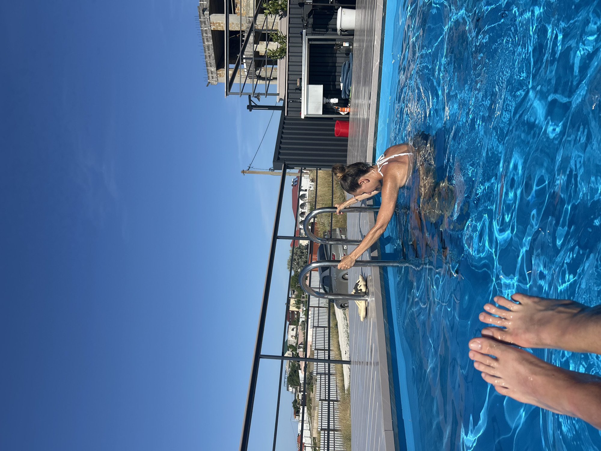 A woman in a white bikini is in a swimming pool, holding onto the ladder. Her head is tilted down, and she appears to be emerging from the water. The pool water is a vibrant blue with visible ripples and reflections. In the foreground, two feet are visible at the surface of the water, suggesting another person is in the pool or near the edge.
Behind the pool, there's a modern structure that looks like it's under construction, featuring exposed concrete and metal beams. Further back, a dark grey shipping container-like building is visible, with some industrial equipment like hoses and a red bucket on it. Beyond these structures, a fence separates the pool area from a landscape with buildings and vegetation, indicating a populated area. The sky is a clear, bright blue, suggesting it is daytime and the weather is sunny. The location context is Мысовское сельское поселение, Ukraine. There is no visible text in the image.