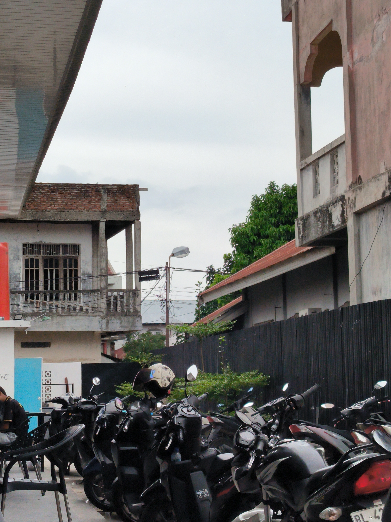 The image depicts a street scene in Lhokseumawe, Indonesia. A row of parked motorcycles dominates the foreground. A helmet rests on one of the motorcycles. A person sits in a chair near the motorcycles. Buildings of varying styles and conditions form the backdrop. Some buildings show signs of aging. Trees and a fence intermix with the architectural elements. Overhead power lines are visible. The sky is overcast.
