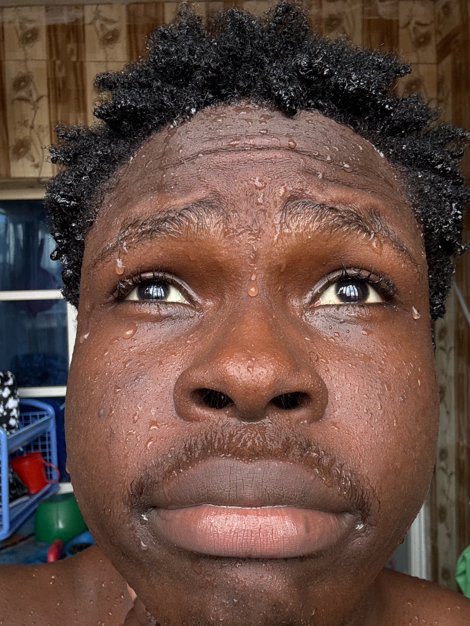 This is a close-up, eye-level, portrait-style photograph of a person in Ihiagwa, Nigeria, with visible water droplets on their face and hair. The subject's dark, curly hair is wet, and numerous water droplets are scattered across their forehead, cheeks, and chin. Their eyes are open and looking upwards, and their lips are slightly parted. The skin appears slightly shiny due to the moisture. The background is softly blurred but suggests an indoor environment with what appears to be tiled walls and some shelving or storage units with colorful objects, including a blue basket and a red bucket. The lighting suggests it may be daytime.