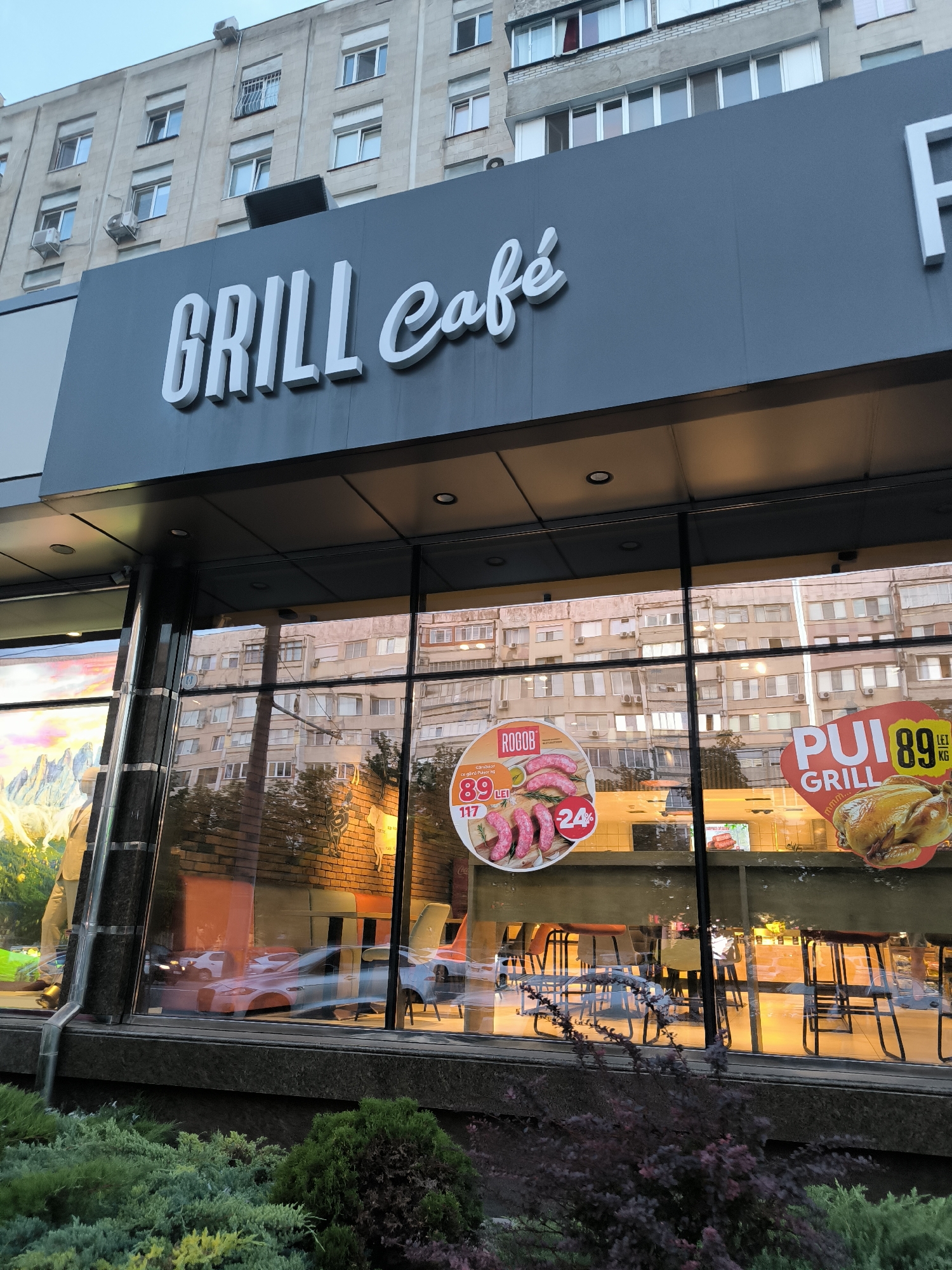 The image shows the exterior of a "Grill Cafe" establishment in Chișinău, Moldova. The cafe has large glass windows displaying various food promotions. One promotion features sausages with a price of 89 lei and "24%". Another promotion shows a whole grilled chicken with the text "PUI GRILL" and a price of 89 lei per kg. Inside the cafe, tables and chairs are visible, as well as reflections of the buildings opposite. The building housing the cafe appears to be a multi-story residential building. The foreground shows some greenery and bushes. The sky is overcast.