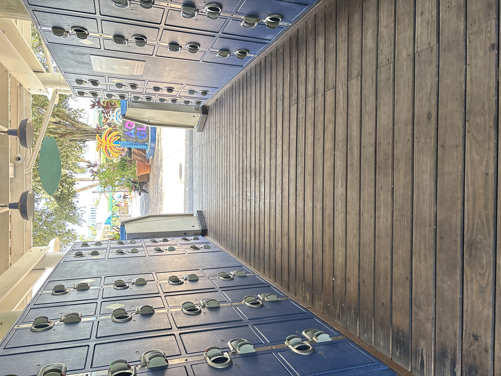 This image captures a view of an outdoor locker area, likely at an amusement park or water park, with a boardwalk in the foreground. The lockers, which are dark blue, are arranged in rows on either side of a wooden pathway. Each locker has a silver handle mechanism. In the background, beyond the lockers, there is a glimpse of a sunny outdoor environment with trees, plants, and some structures that appear to be part of a recreational facility. There are no people clearly visible in the immediate vicinity of the lockers. The location is identified as being in Tampa, United States of America.