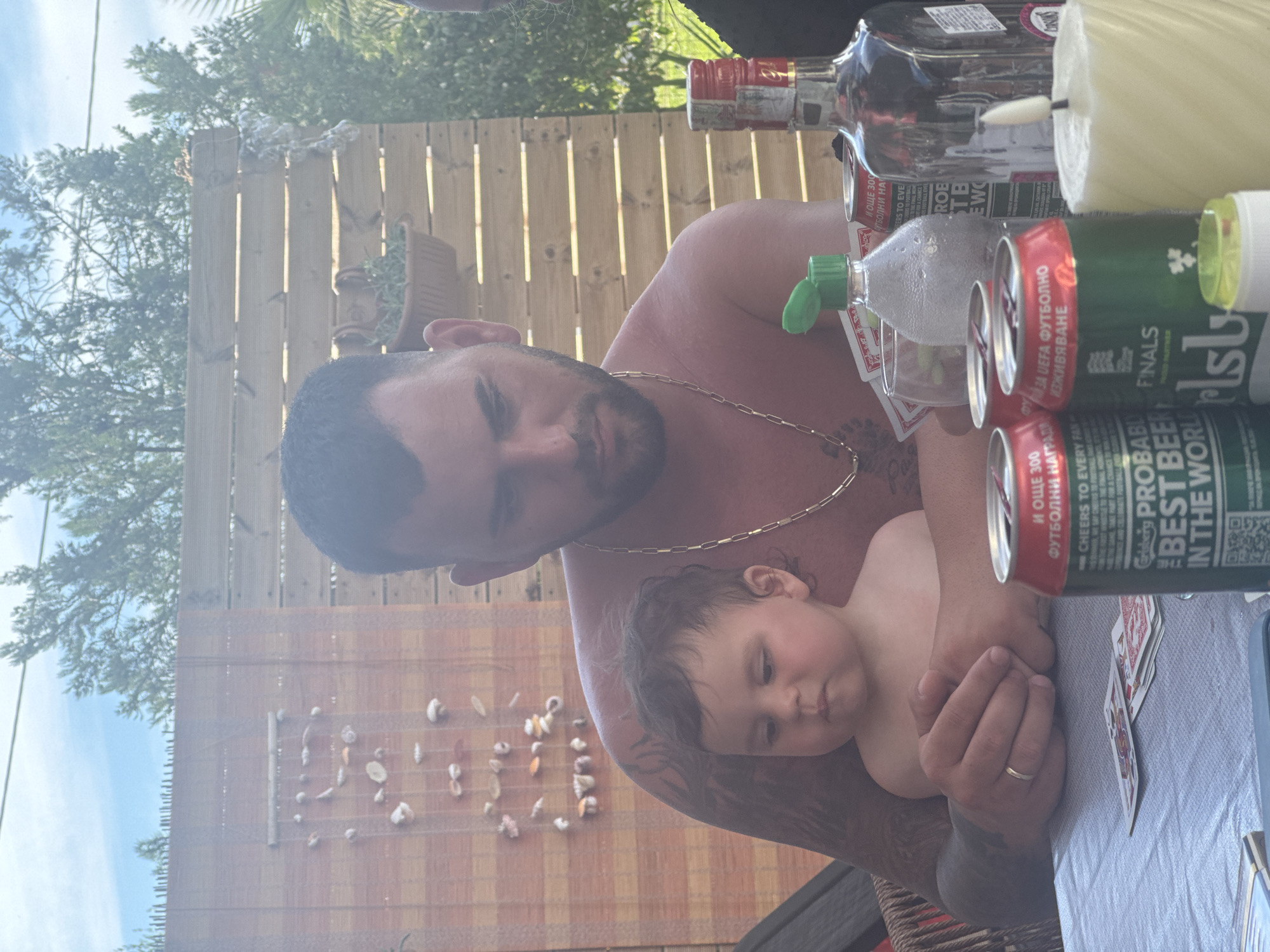 A man is holding a baby on his lap. They are seated at a table, outdoors, with a wooden fence in the background. The man has tattoos on his arm and is wearing a gold chain. The baby is looking down. On the table are several cans of Carlsberg beer, a bottle of alcohol, a candle, and a deck of cards. The scene appears to be in Pallini Municipal Unit, Greece, judging by the text on one of the Carlsberg cans.