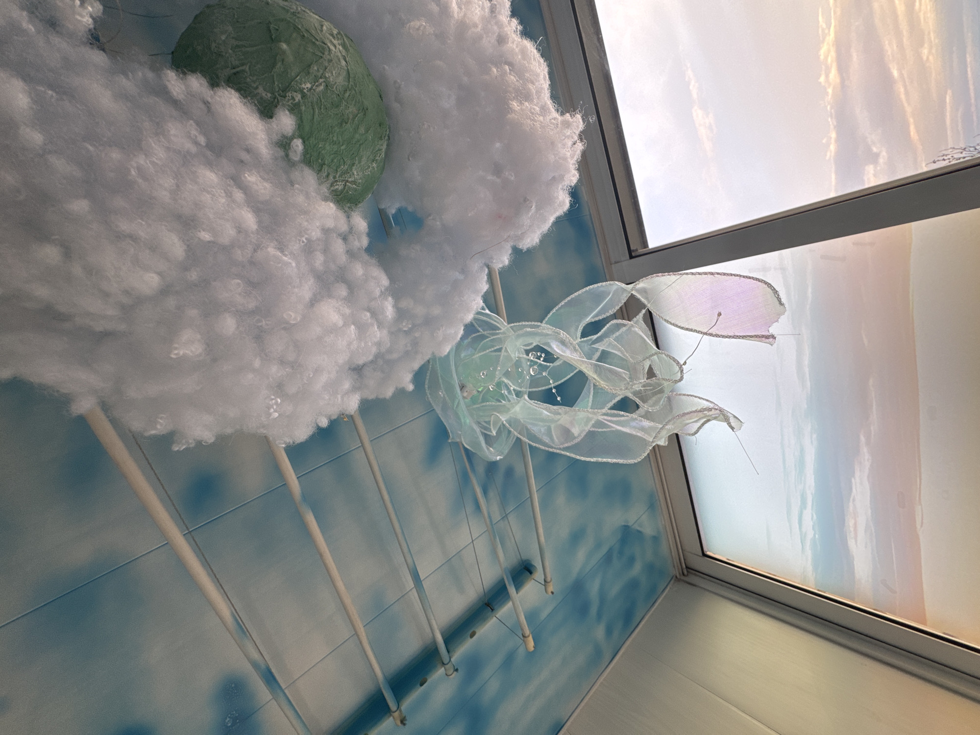 The image shows an indoor scene with a whimsical decoration hanging near a window. The decoration consists of white, fluffy material resembling clouds, a green, rounded object possibly representing a planet or landmass, and a translucent, iridescent ribbon sculpture hanging below the "clouds". The decoration is suspended by multiple white rods. The wall behind the decoration is tiled in a blue and white pattern. The window is partially visible, with a view of the sky at sunset or sunrise. The exact location in Novo-Nikol'skoe, Russia cannot be determined from the image. There are no people visible in the image.
