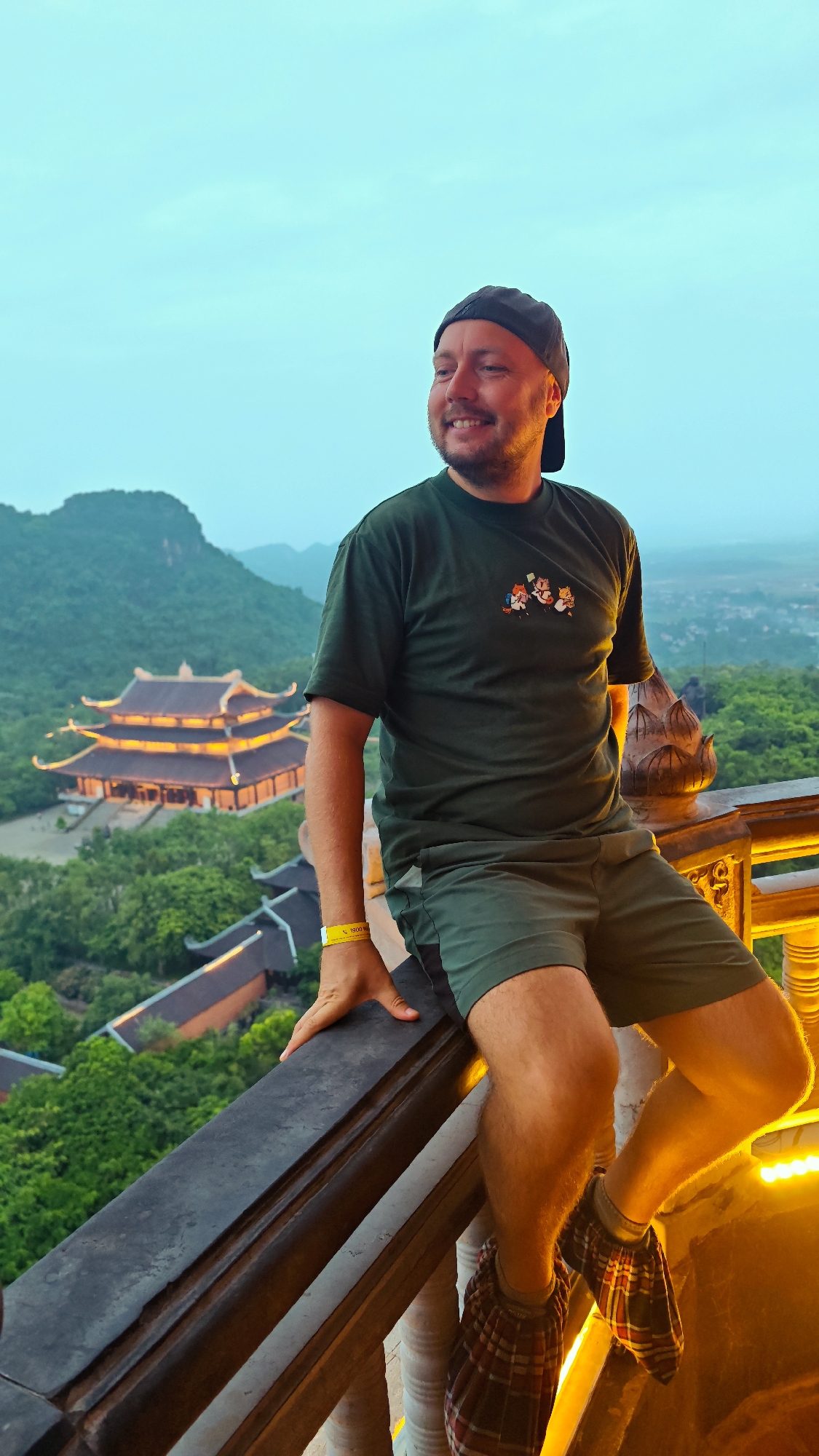 A man is perched on a railing overlooking a valley with a Buddhist temple. The man is wearing a dark green t-shirt with a cartoon graphic, green shorts, a black baseball cap, and plaid shoe covers. He is smiling and looking to his left. The temple, illuminated with warm lights, has a multi-tiered roof and is surrounded by lush green trees. In the background, mountains are visible under a clear, pale blue sky. The railing is made of dark wood with ornate carvings, and a bright yellow wristband is visible on the man's left wrist. The scene is likely in the vicinity of Tây Hoa Lư Ward, Vietnam, given the architectural style of the temple and the surrounding landscape.