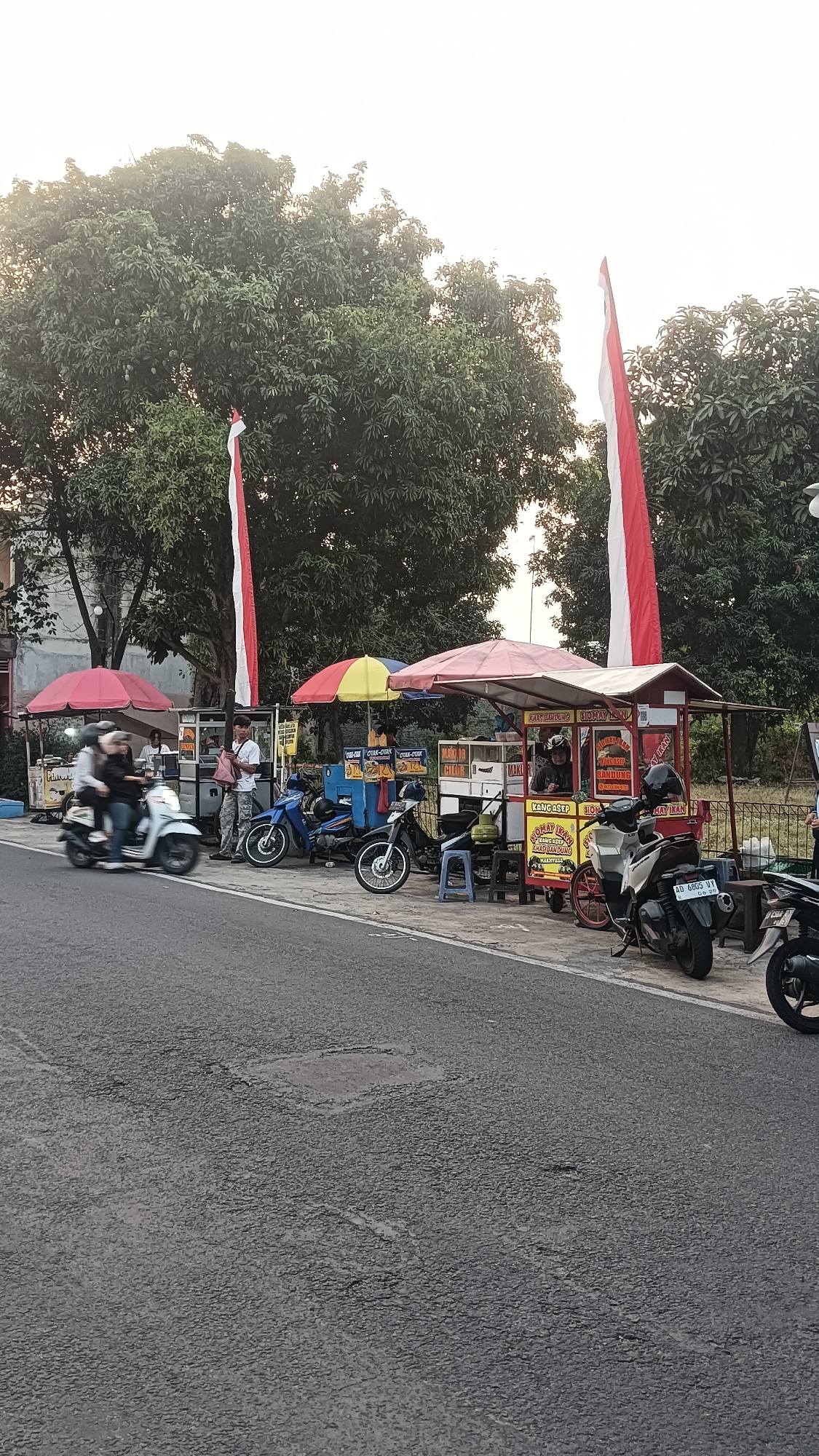 The image depicts a street scene in Sragen Tengah, Indonesia, likely during daylight hours given the lighting. In the foreground, a gray asphalt road with visible patches and some texture is present. On the right side of the road, several motorcycles are parked along a sidewalk. One motorcycle in the immediate foreground has a license plate readable as "AD 6805 VY".
Adjacent to the sidewalk are several food stalls, some with red and white Indonesian flags. These stalls appear to be selling food, with signage visible in what is likely Indonesian. One stall has a red and yellow umbrella, and another has a red canopy with a more elaborate structure. People are visible at and around the stalls.
On the left side of the image, a motorcycle with three occupants is in motion, blurring due to movement. Further to the left, there are more food stalls, one with a red umbrella. Trees with dense green foliage line the background, suggesting a park or green space behind the stalls. The sky is bright, indicating daylight, possibly late afternoon or early morning given the soft light. The overall scene suggests a bustling roadside market or food vendor area.