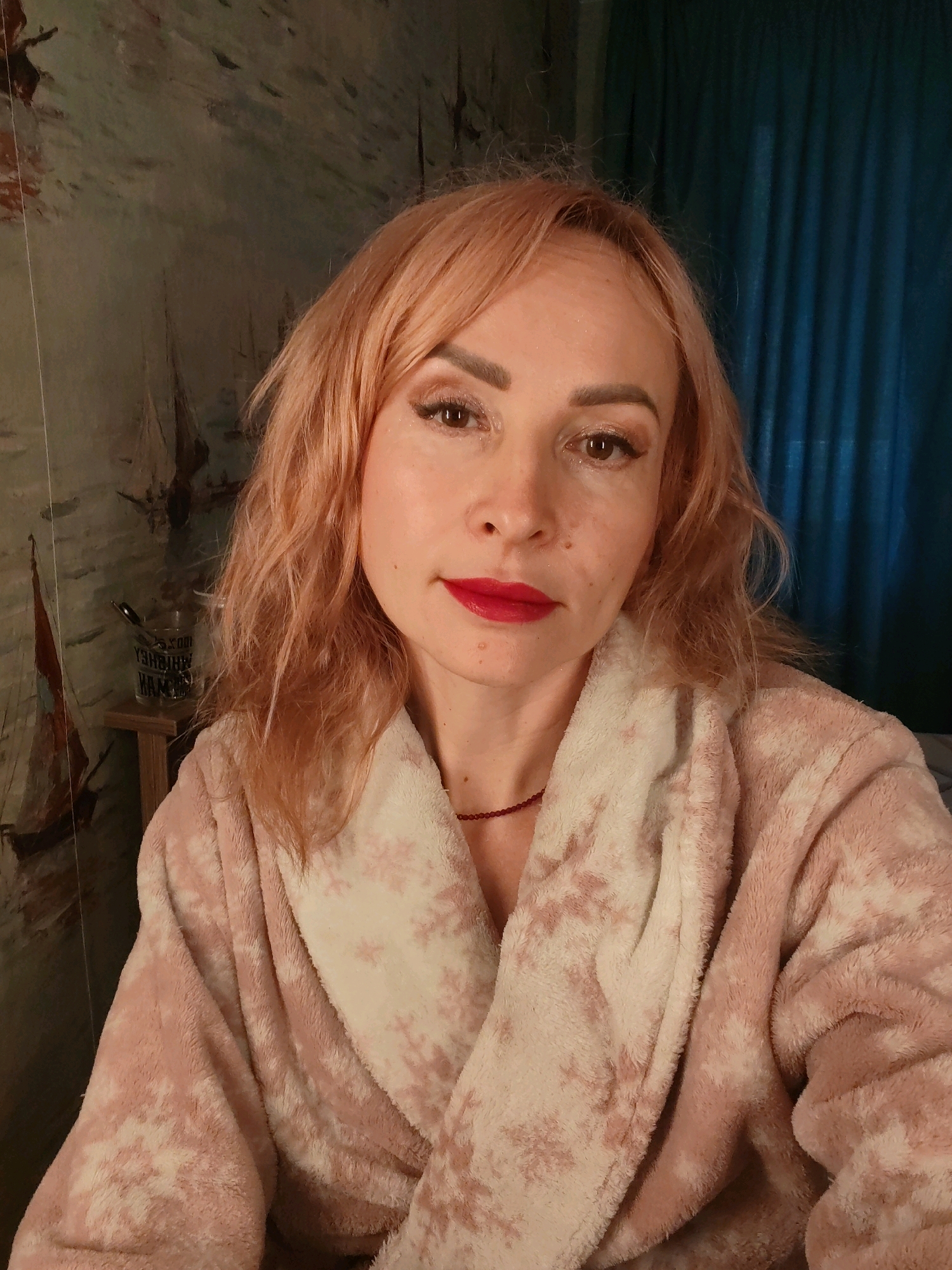 A middle-aged woman with peachy-pink hair and fair skin is positioned centrally in the frame, looking directly at the camera. She is wearing a pink and white patterned bathrobe. Her makeup includes bold red lipstick and shimmering eyeshadow. Behind her, the background features a wall with a mural of sailboats and a dark blue curtain. To her left, a small wooden shelf holds a glass with pens or pencils. The lighting is warm and somewhat dim, suggesting an indoor setting. There is no specific indication of the city of Chaikovsky, Russia, in the image itself.