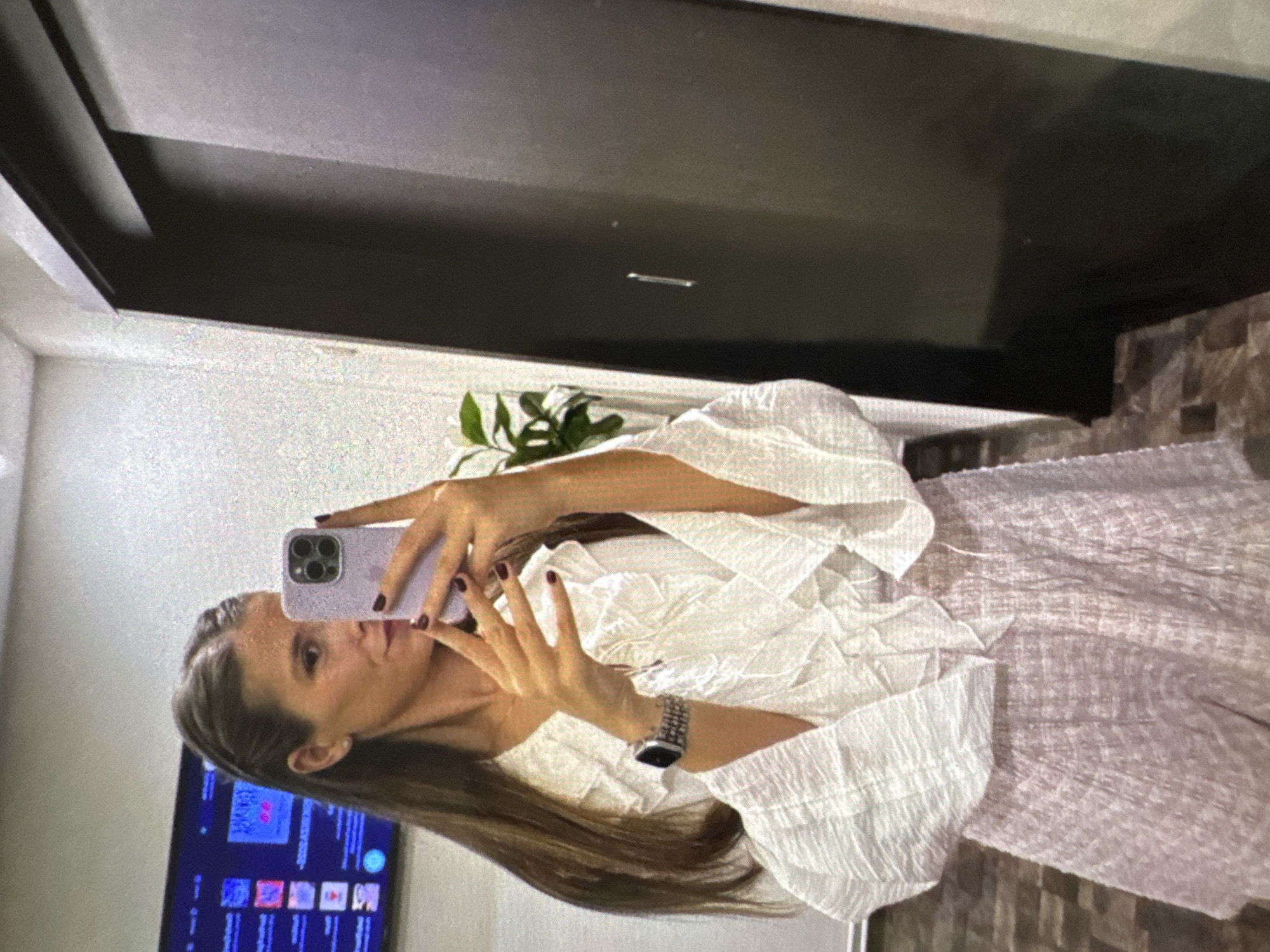 A female individual is captured in a mirror selfie, taken indoors. She has long brown hair, parted down the middle with two small braids pulled back from the front. Her face is partially obscured by a light purple iPhone, held in her right hand, which displays a triple-lens camera array. Her fingernails are painted a deep burgundy. She wears a smartwatch with a metallic mesh band on her left wrist. Her attire consists of a white, crinkled or textured blouse with ruffled details around the V-neckline and wide, bell-shaped sleeves, paired with light pink or lavender textured shorts.
The background environment features a light-colored, speckled wall. A wall-mounted television or monitor displays various colorful application icons or video thumbnails. To the right, a dark, possibly sliding, door or panel is visible with a subtle vertical handle and an overhead track. A green houseplant is partially visible behind the subject's right arm. The floor is composed of patterned, rectangular tiles in shades of brown and grey. The specific location within Vratsa, Bulgaria, cannot be visually identified from the image content.