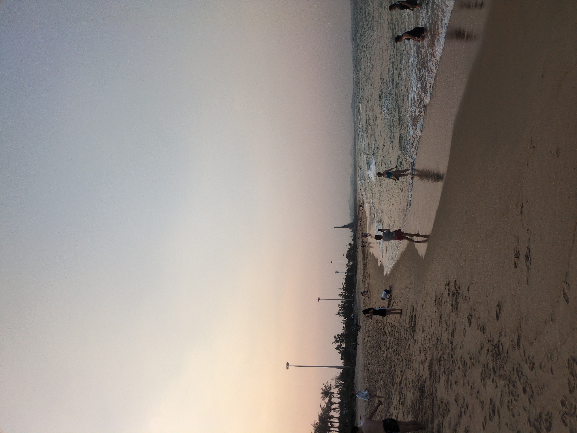 The image depicts a beach scene at sunset in Tuy Hòa, Vietnam. The sky is a gradient from light grey to orange near the horizon. The beach is sandy and textured with numerous footprints. Several people are present, some wading in the water at the edge of the waves, others standing or walking along the shore. Streetlights are visible along the beach. The water is slightly choppy with small waves.