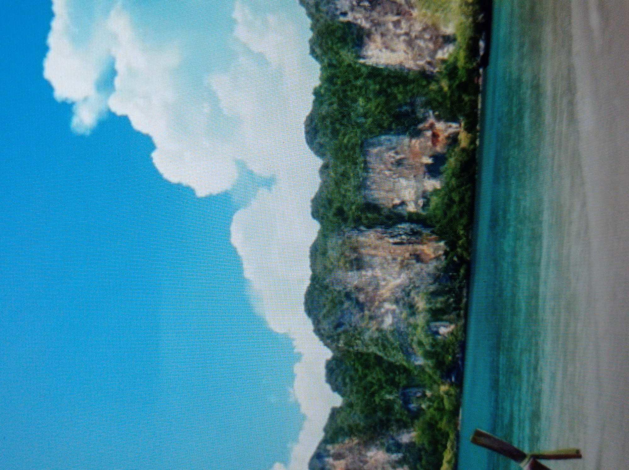 The image displays a tropical beach scene with turquoise water, a sandy shore, and lush green cliffs dotted with rock formations under a bright blue sky with fluffy white clouds. A wooden post, possibly part of a pier or a sign, juts out from the water on the left side of the frame. The scene does not depict any elements related to the city of Mukachevo, Ukraine.