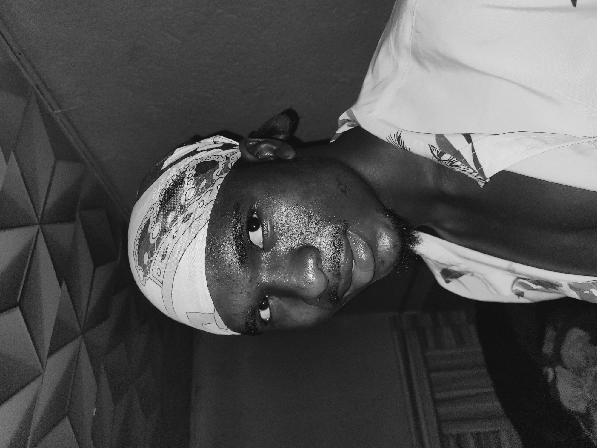 In a black and white photograph, a young Black male is shown from the chest up, looking directly at the camera. He is wearing a white bandana on his head with a patterned design and a white collared shirt with a similar pattern along the collar. His skin appears to be somewhat oily. The background consists of a textured ceiling and a wall. The lighting is somewhat dim, casting shadows. The individual's expression is neutral. The setting is indoors, likely a residence. No specific urban landmarks or environmental features are visible to pinpoint a location within Bukuru, Nigeria.