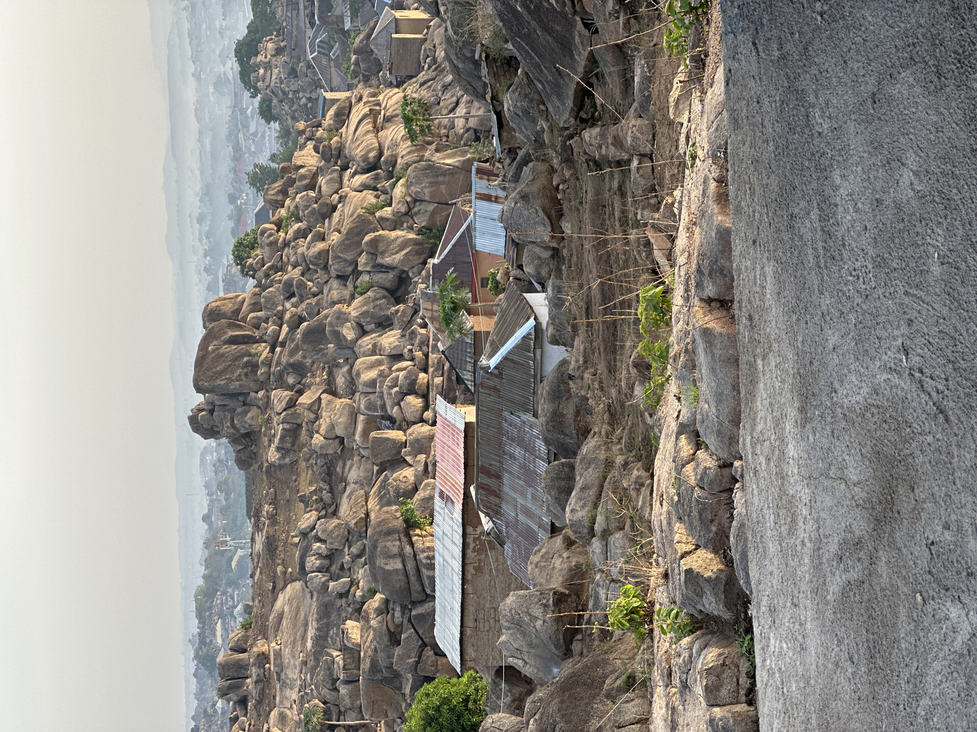 The image presents a rugged, rocky landscape featuring a settlement nestled among prominent boulders, likely on a hillside or mountain. In the foreground, a large, relatively flat grey rock surface occupies the right side, with sparse green plants and dry branches growing along its edge.
The midground is dominated by numerous large, rounded boulders, varying in size and color from grey to earthy brown, piled and scattered across the terrain. Integrated within this rocky environment are several single-story houses or buildings. These structures are characterized by their corrugated metal roofs, some exhibiting a rusty reddish-brown patina, while others are silver or grey. The walls of the houses, where visible, appear to be constructed from rough stone or earthy materials, blending with the natural rock formations. Scattered green vegetation, including small trees and bushes, dots the rocky landscape, particularly around the dwellings.
In the hazy background, a vast valley or plain stretches out under a light grey sky. Distant structures, suggesting a town or city, are faintly visible through the atmospheric mist. The lighting indicates daytime, possibly late afternoon or early morning, with soft, diffuse sunlight illuminating parts of the scene. No individuals are visible, and there is no discernible text in the image. The scene depicts a community integrated into a challenging natural setting, typical of parts of Jos, Nigeria.
