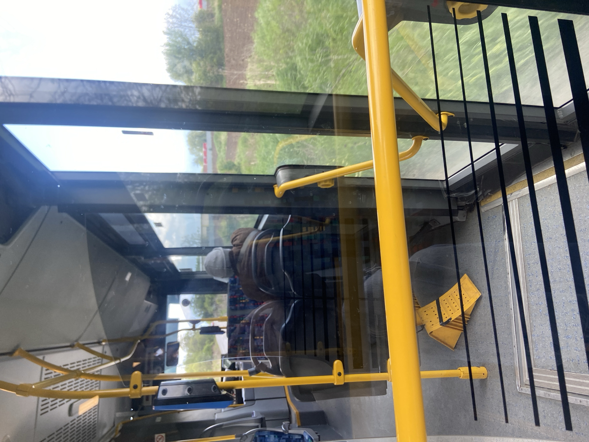 The image, taken inside a bus and rotated 90 degrees clockwise, captures a bright daytime scene from a passenger's perspective, looking forward and out through the windows. The interior of the bus is dominated by multiple bright yellow grab rails and vertical poles, contrasting with the light grey, speckled floor. Dark grey or black seats are visible, both directly inside the bus and as reflections on the windows.
One person, wearing a light-colored cap and a brown jacket, is seated further down the bus aisle, mostly discernible as a reflection on the window. On the bus floor near a window, a distinctive yellow object, possibly a folded ramp or a safety sign with a textured surface and black markings, is visible.
Through the bus windows, a rural or semi-rural landscape is seen. The exterior view features expansive green fields and patches of tilled brown earth under a bright sky. Distant trees with green foliage are visible, indicating clear, bright weather, likely late morning or early afternoon. The windows show significant reflections of the bus's interior, superimposing elements like seats and ceiling onto the outdoor view.
The scene depicts a moment of travel by public transport through what appears to be an agricultural area, consistent with the Zhiten, Bulgaria, context. The light quality suggests good weather. There is no clearly visible text in the image.